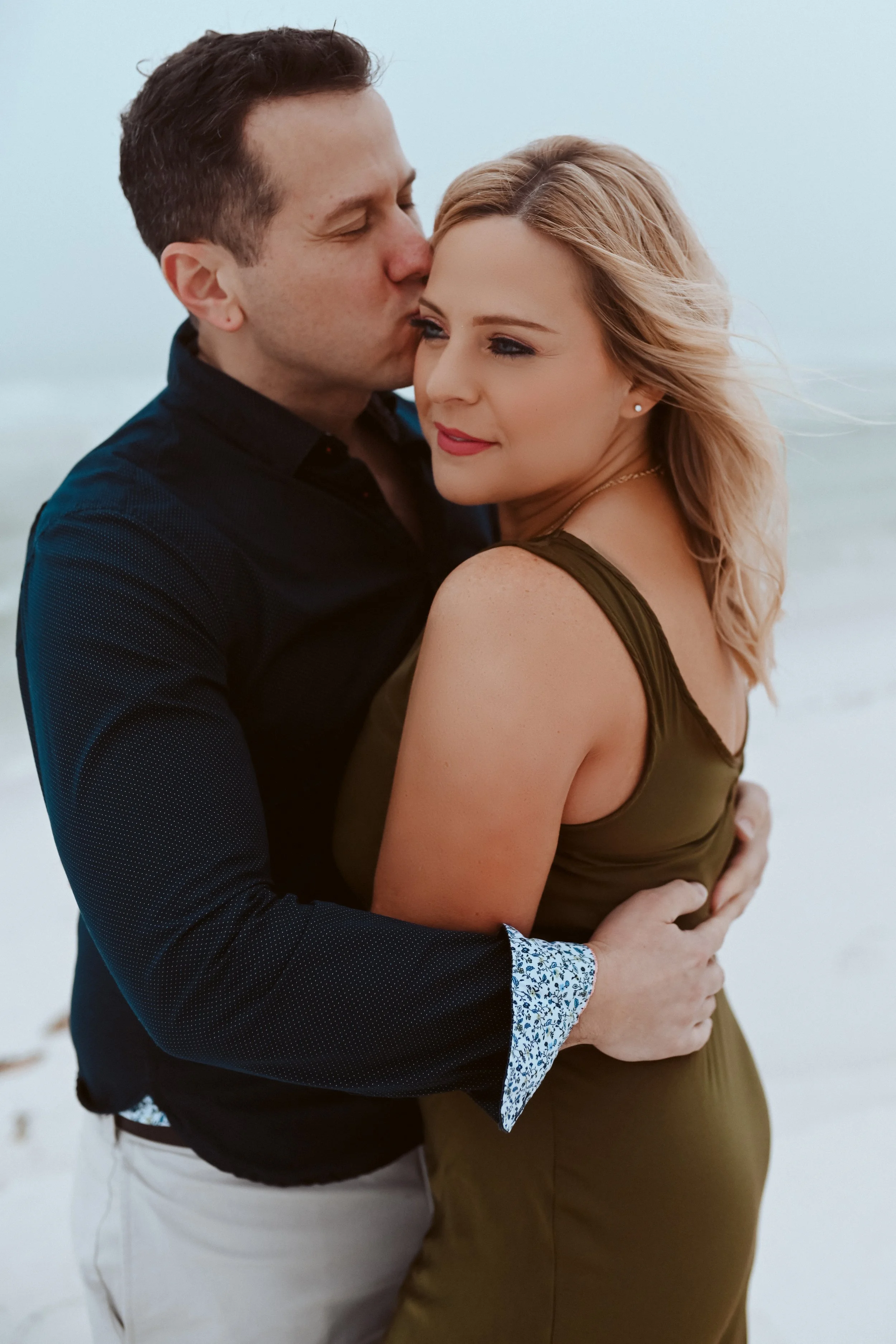A couple embracing on a beach, with the man kissing the woman on the cheek while the woman looks to the side, slightly smiling.