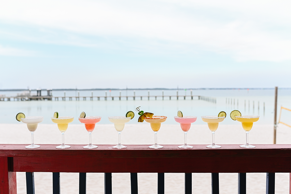 Row of colorful margarita cocktails with lime garnishes on a red wooden bar overlooking a beach and ocean in daytime.