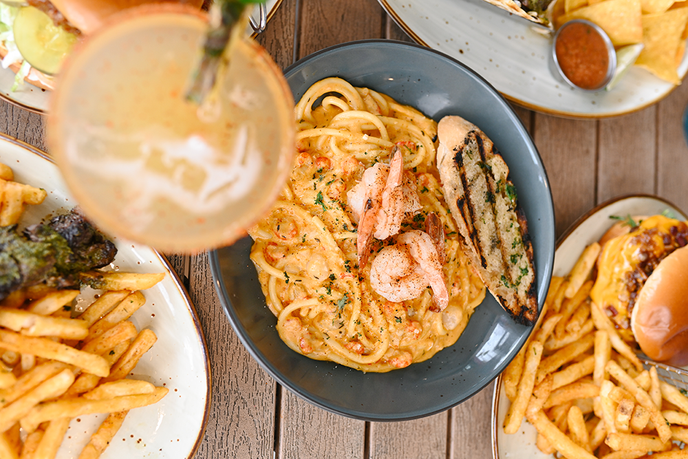 A plate of creamy shrimp pasta with grilled bread, surrounded by other plates of fries and burgers, with a drink in the foreground.