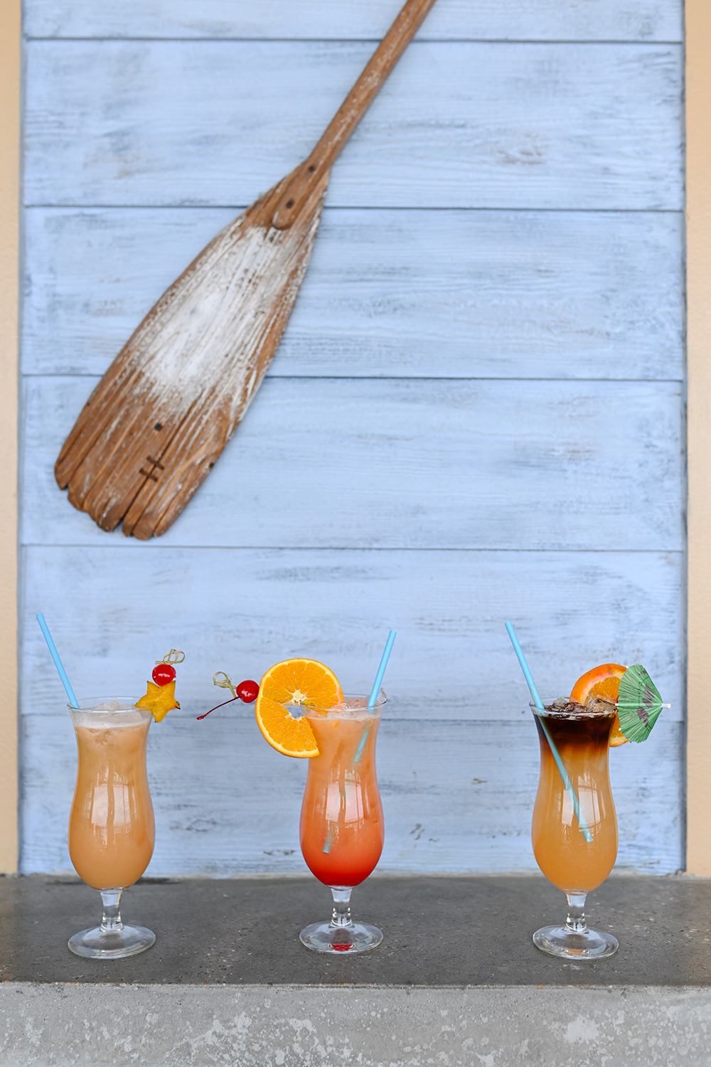 Three tropical cocktails on a counter in front of a weathered light blue wooden wall with a decorative paddle.