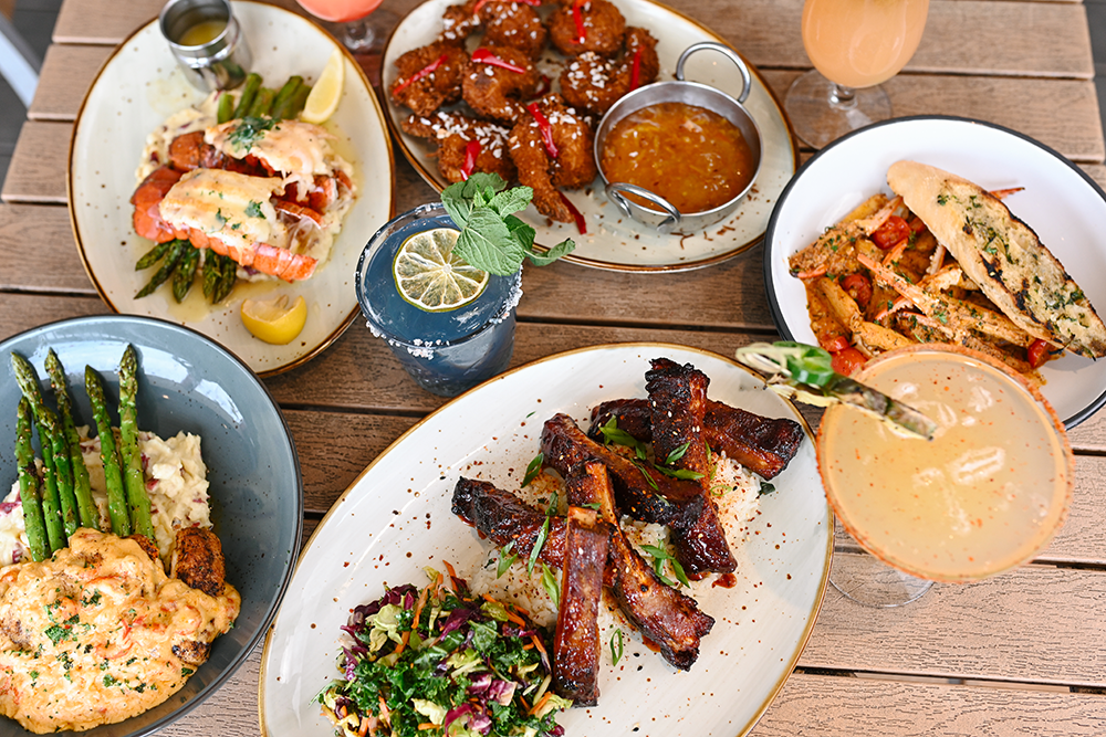 Assorted summer barbecue dishes on a wooden table including grilled chicken and asparagus, ribs, fried chicken bites, and seafood pasta, along with cocktails and sauces.