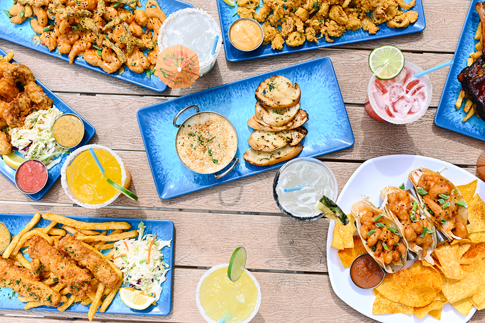 Variety of Tex-Mex dishes including fried chicken, quesadillas, tacos, fried shrimp, chips with salsa, dips, and assorted drinks on wooden table.