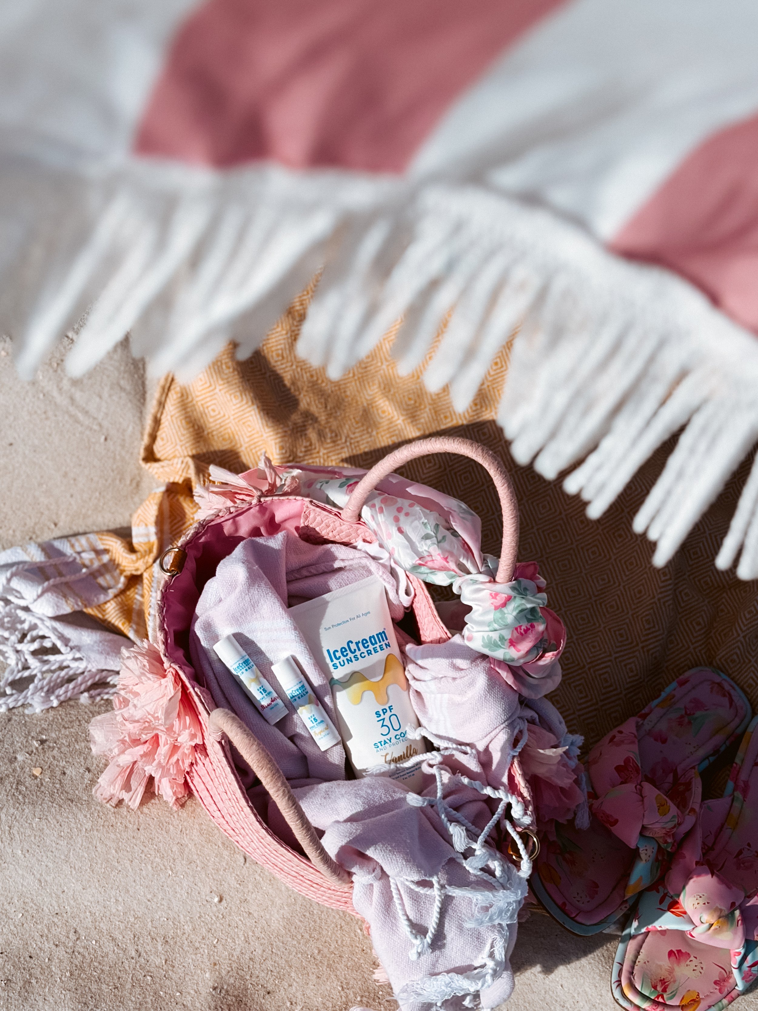 Beach scene showing a pink woven bag filled with sunscreen, covered with light pink and floral fabrics, with a fringed white towel and floral pouch nearby, on sandy ground.