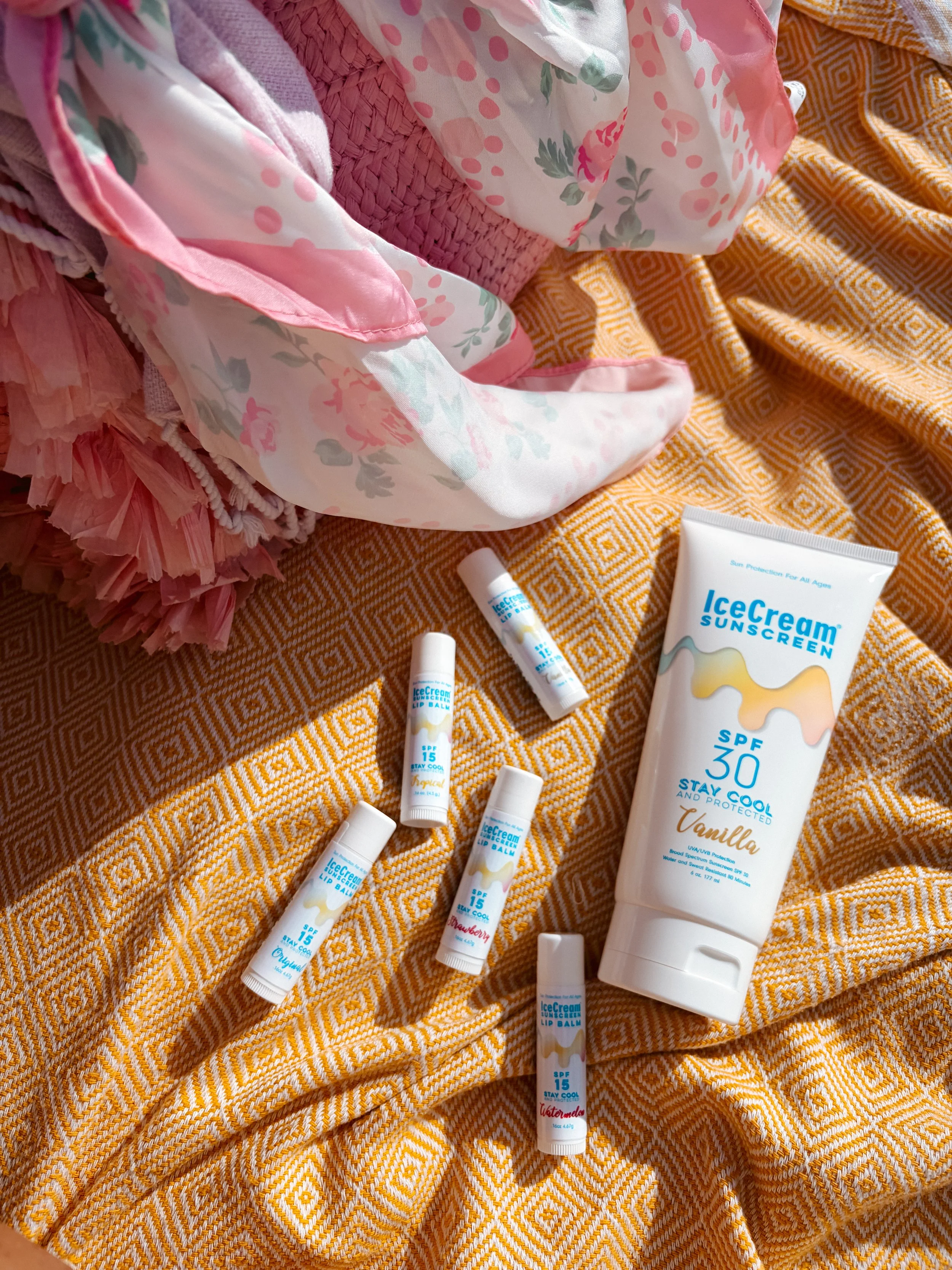 Multiple tubes of IceCream brand SPF 15 lip balm and a tube of IceCream sunscreen SPF 30 in a flat lay photography on a yellow patterned fabric, with pink fabric and a floral-printed cloth nearby.