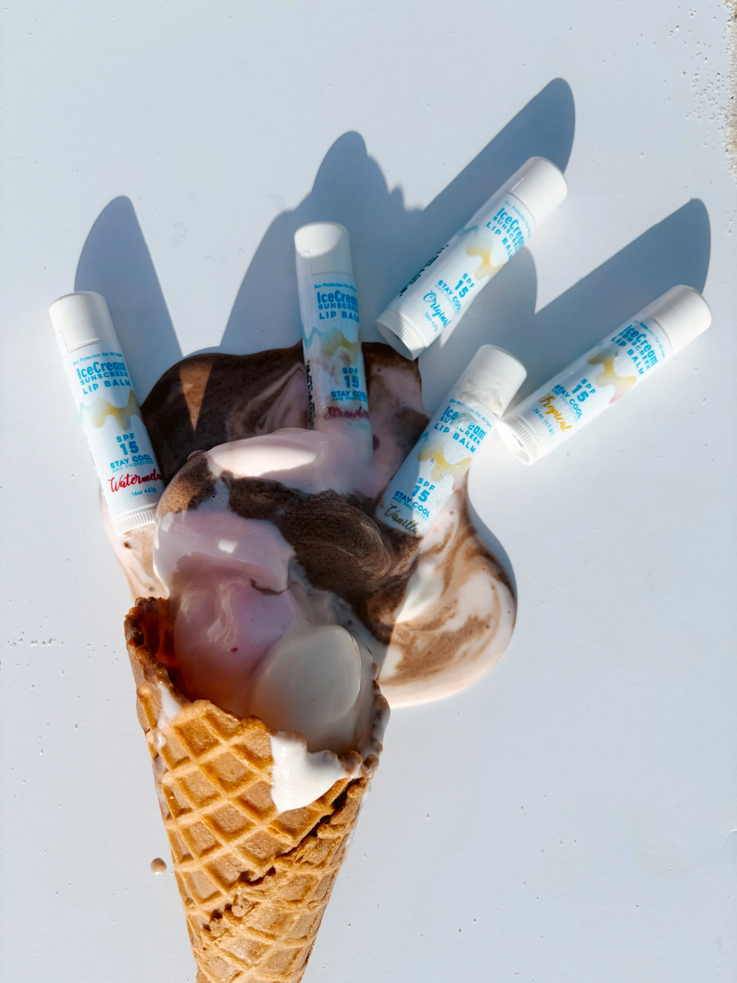 Ice cream cone with several lip balm sticks arranged around it, on a white surface with shadows.