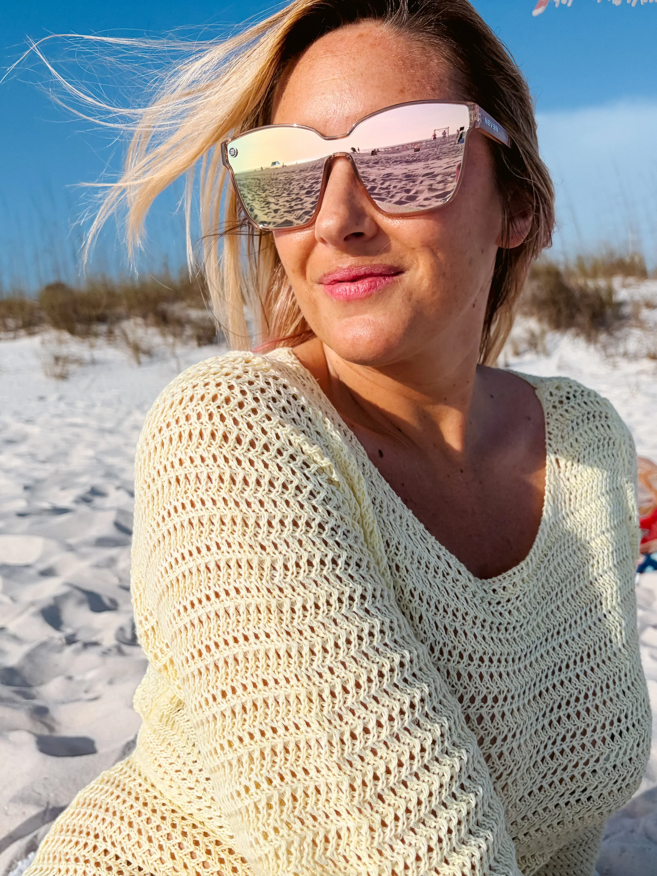A woman sitting on the beach wearing reflective sunglasses and a yellow knitted sweater, with sandy dunes and bushes in the background.