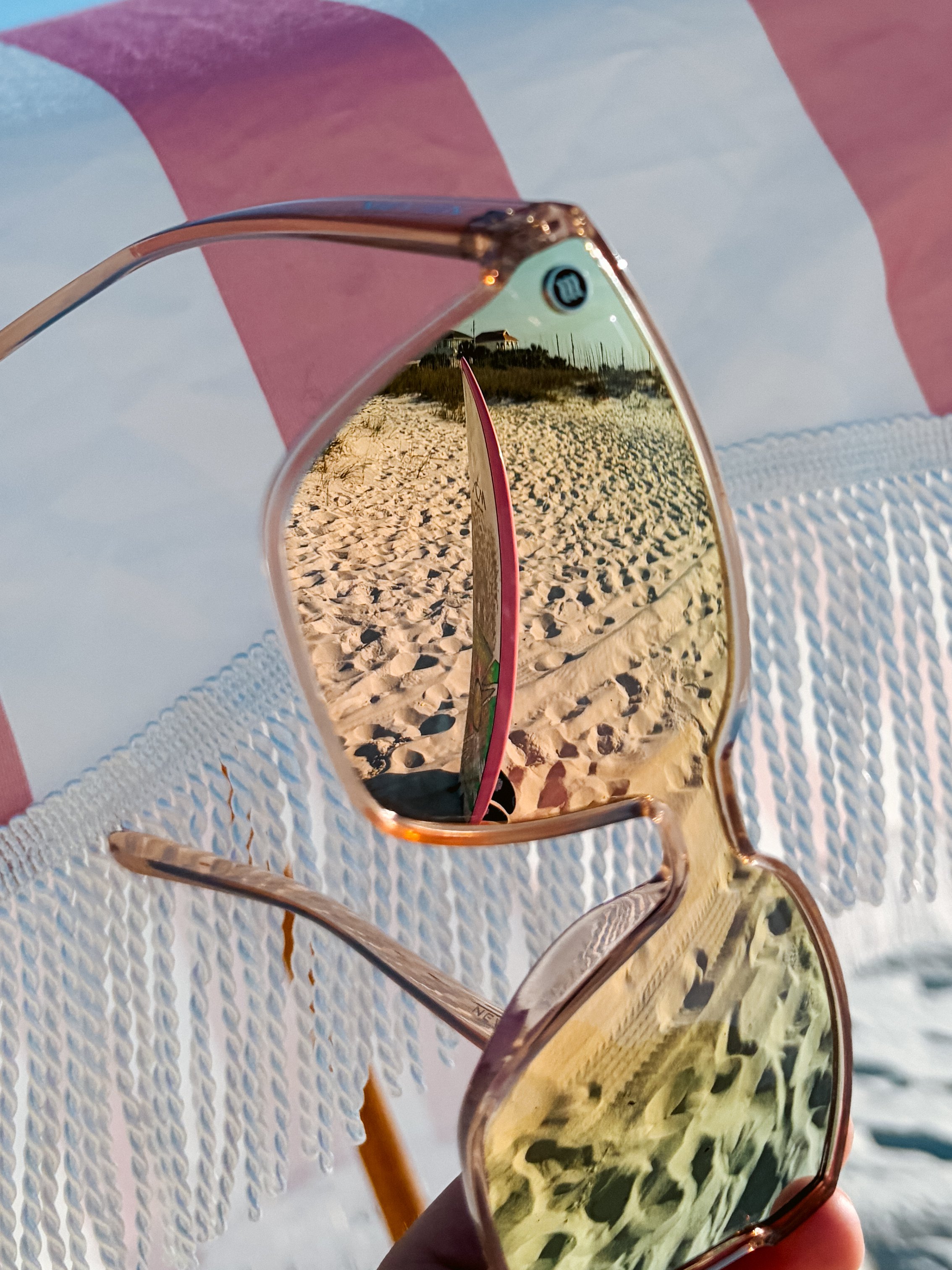 A reflection of a beach scene with sand, a surfboard, and a building in sunglasses lenses held up against the background of a beach chair and white fabric.