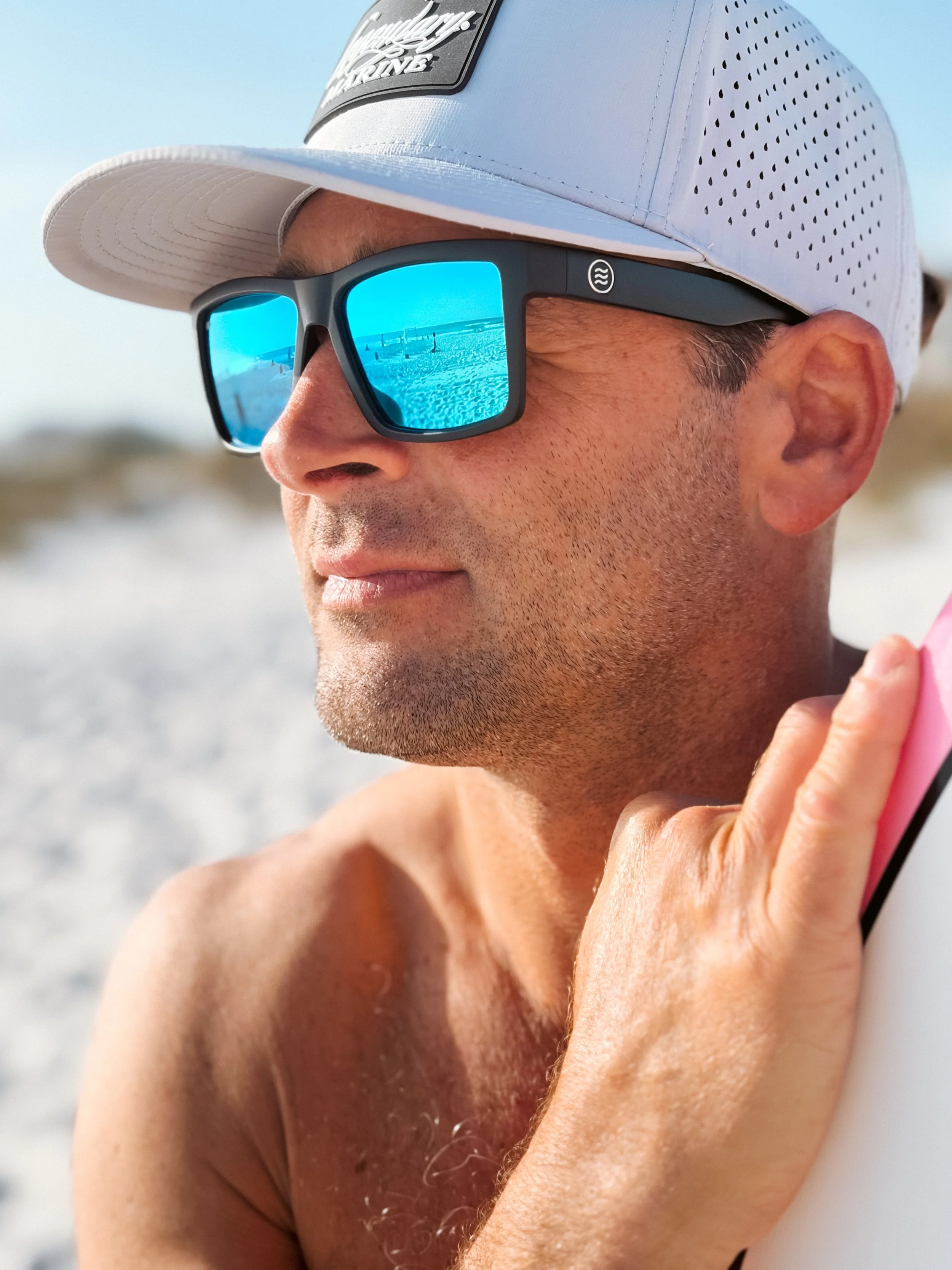 Close-up of a man wearing a white baseball cap and reflective blue sunglasses at the beach, holding a surfboard.