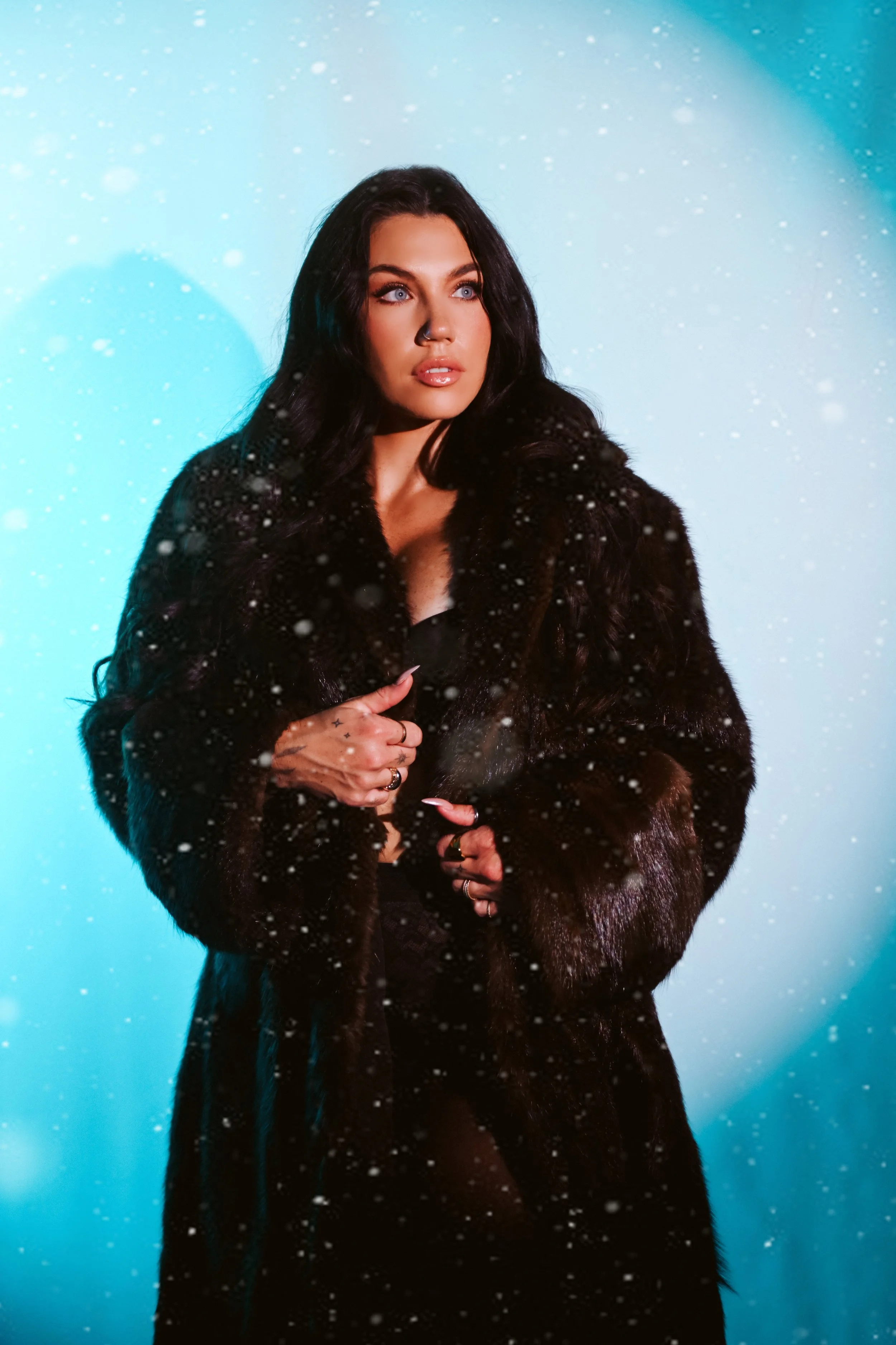 A woman with long dark hair and fair skin, dressed in a black fur coat, standing outdoors in falling snow with a blue background and lit from the side.