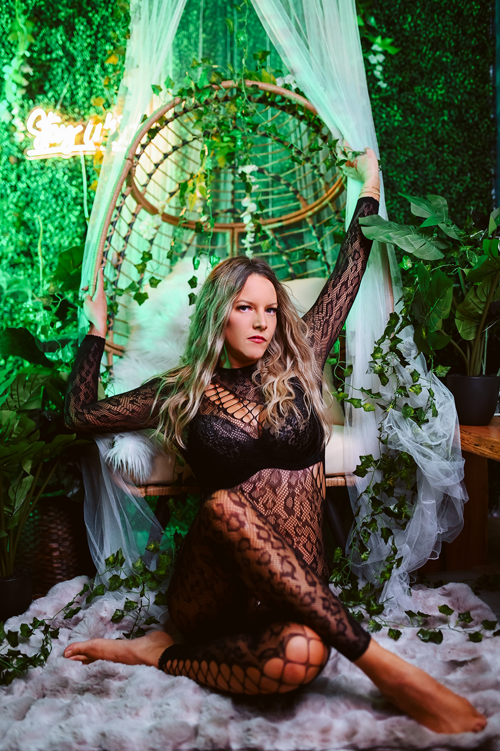 Woman in black lace bodysuit with fishnet pattern posing on fur rug surrounded by green plants in a lush, nature-inspired setting with a hanging canopy and pillows.