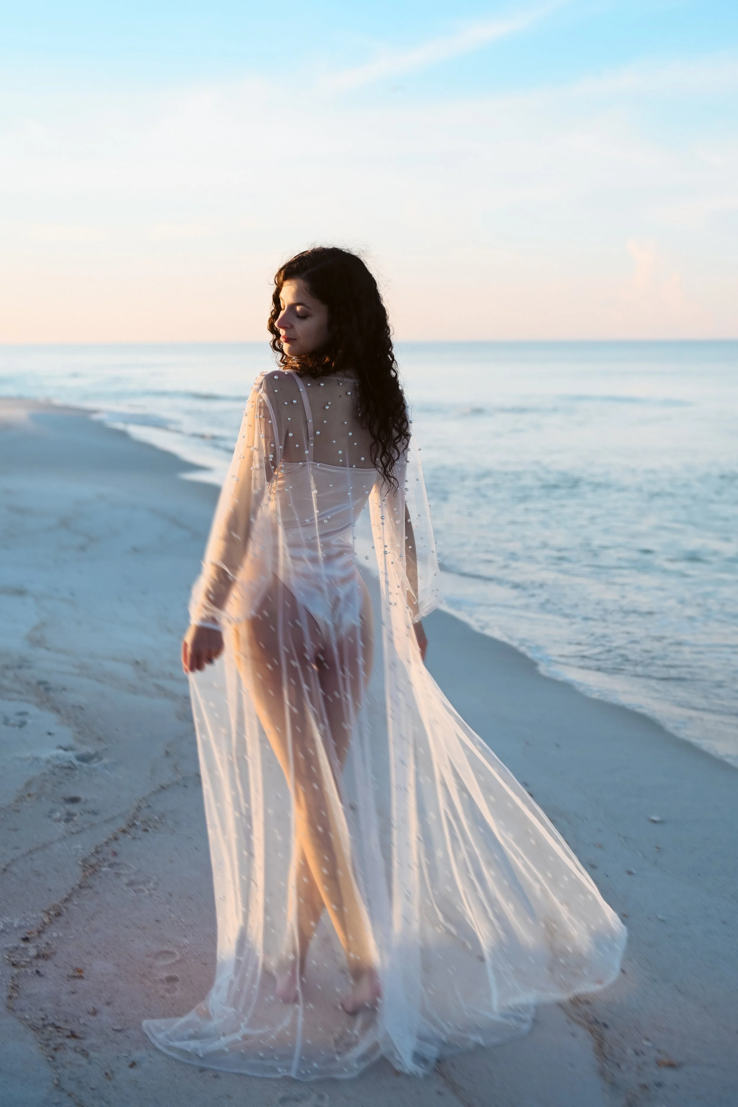 A woman in a sheer, long, pearl-embellished cover-up walking on a sandy beach at sunset.