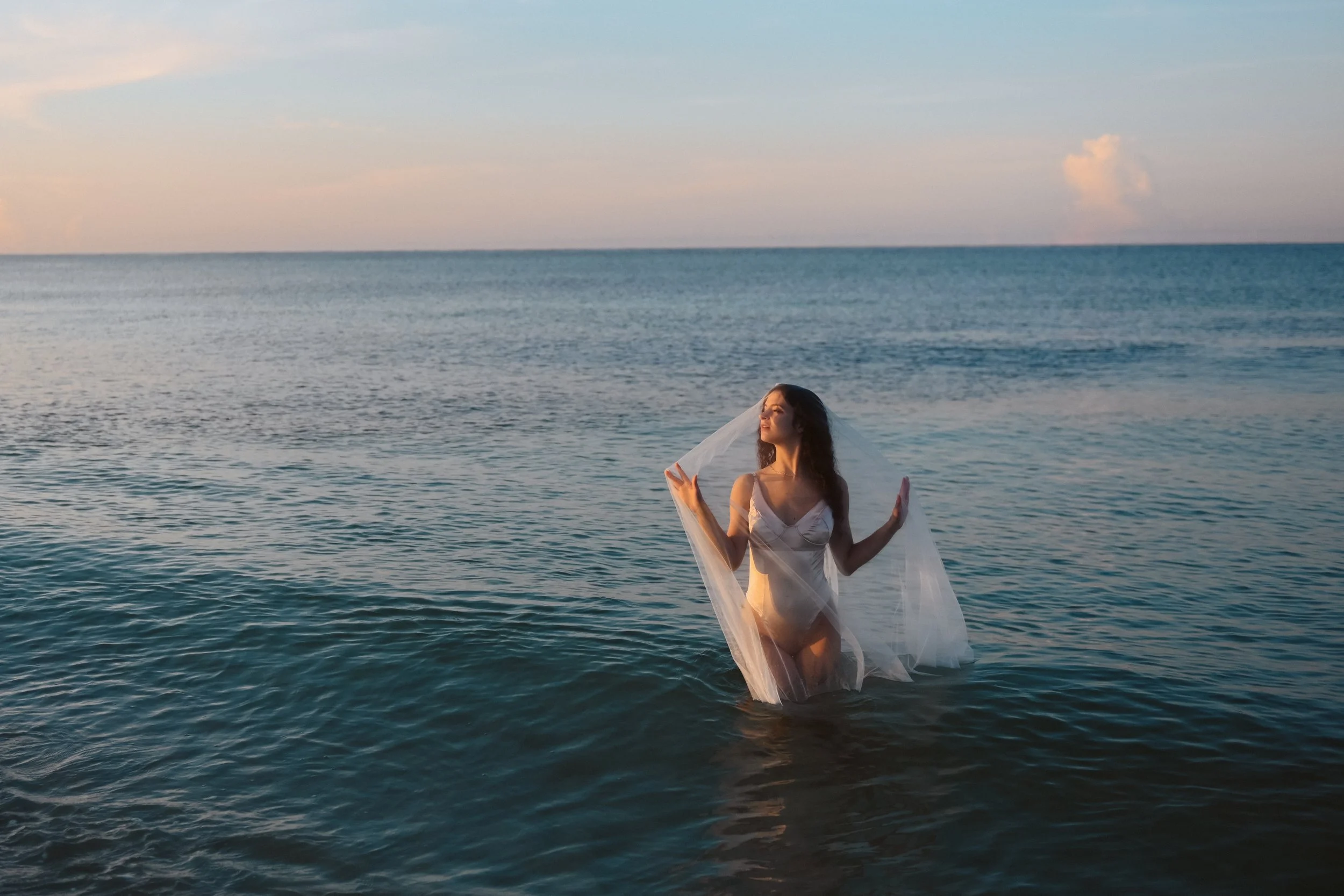 A woman in a white dress kneeling in the ocean during sunset, holding a sheer veil, with the sea extending to the horizon.