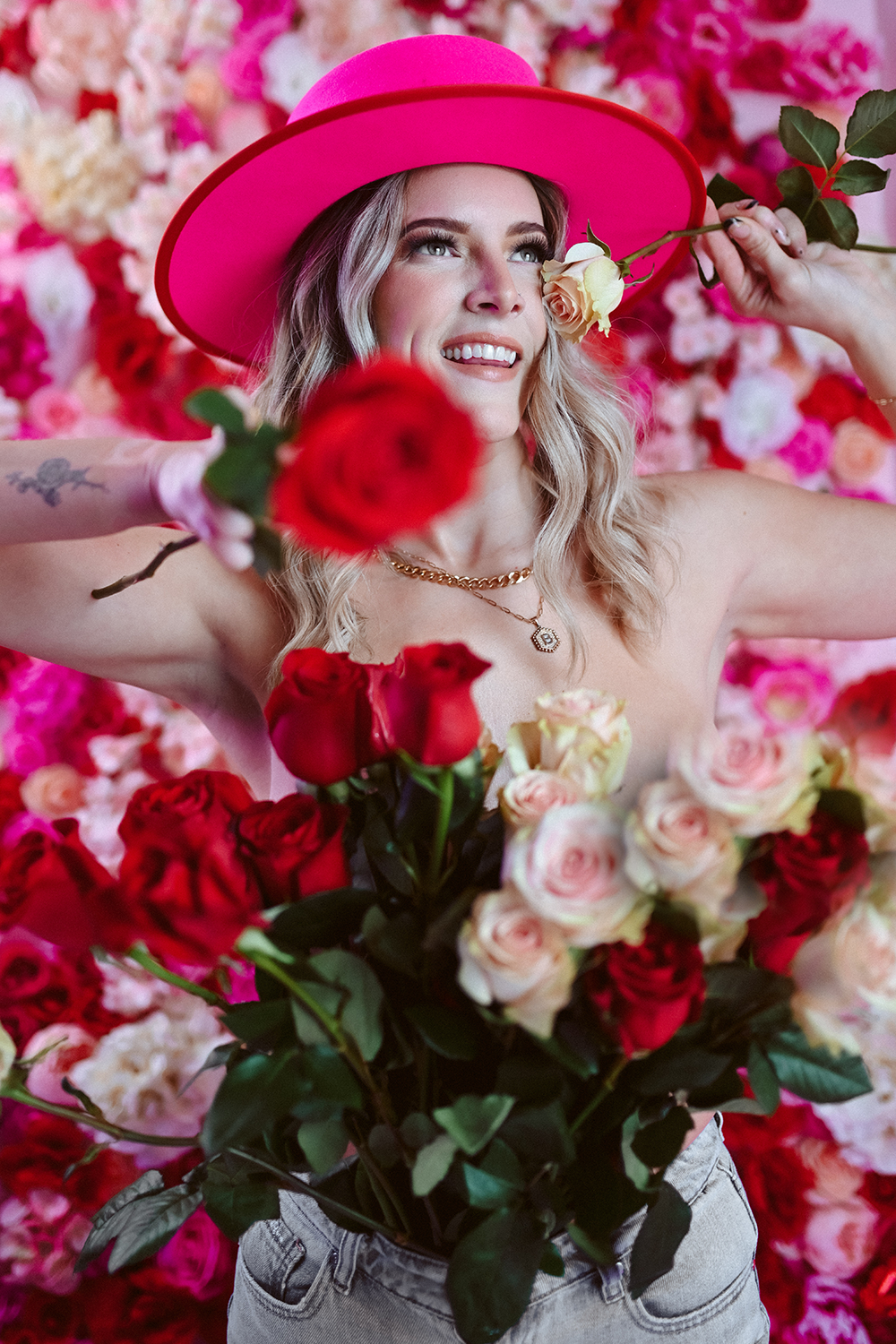 A woman with long blonde hair wearing a pink hat is holding a yellow rose near her face, surrounded by a backdrop of pink, white, and red roses, and also holding a bouquet of red and white roses.