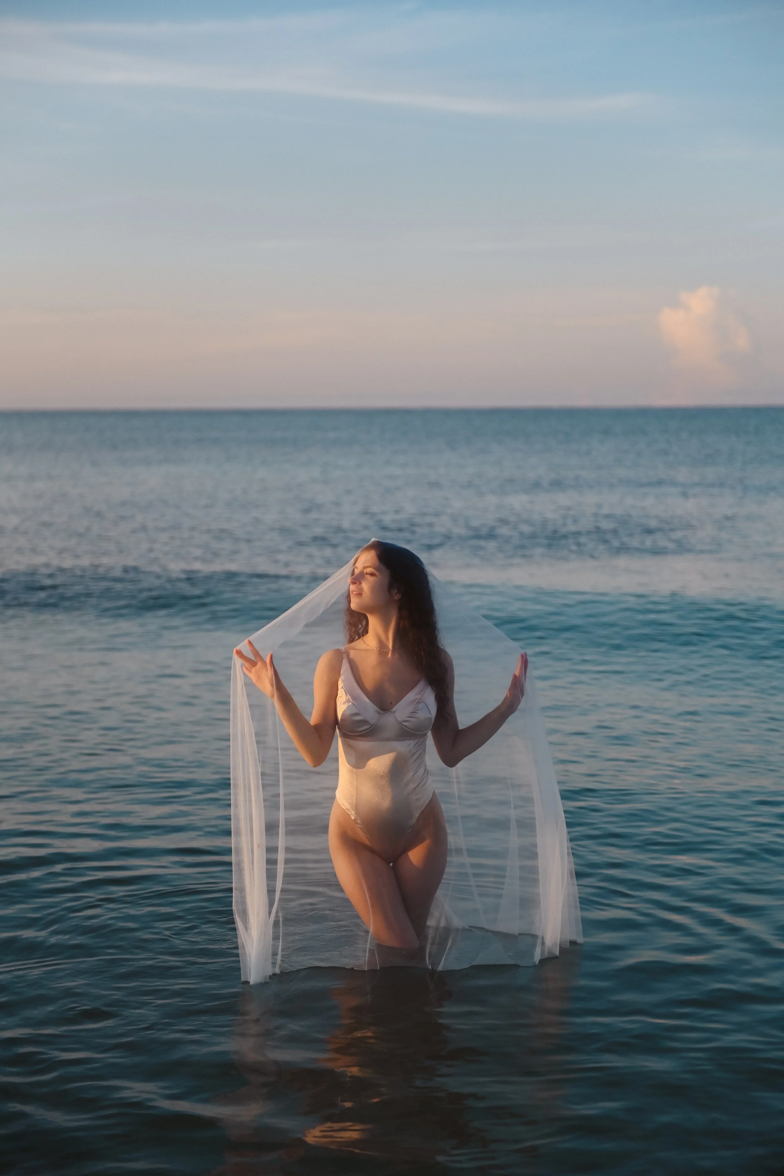 A woman in a beige satin dress standing in the ocean holding a sheer white veil, with the sunset sky in the background.