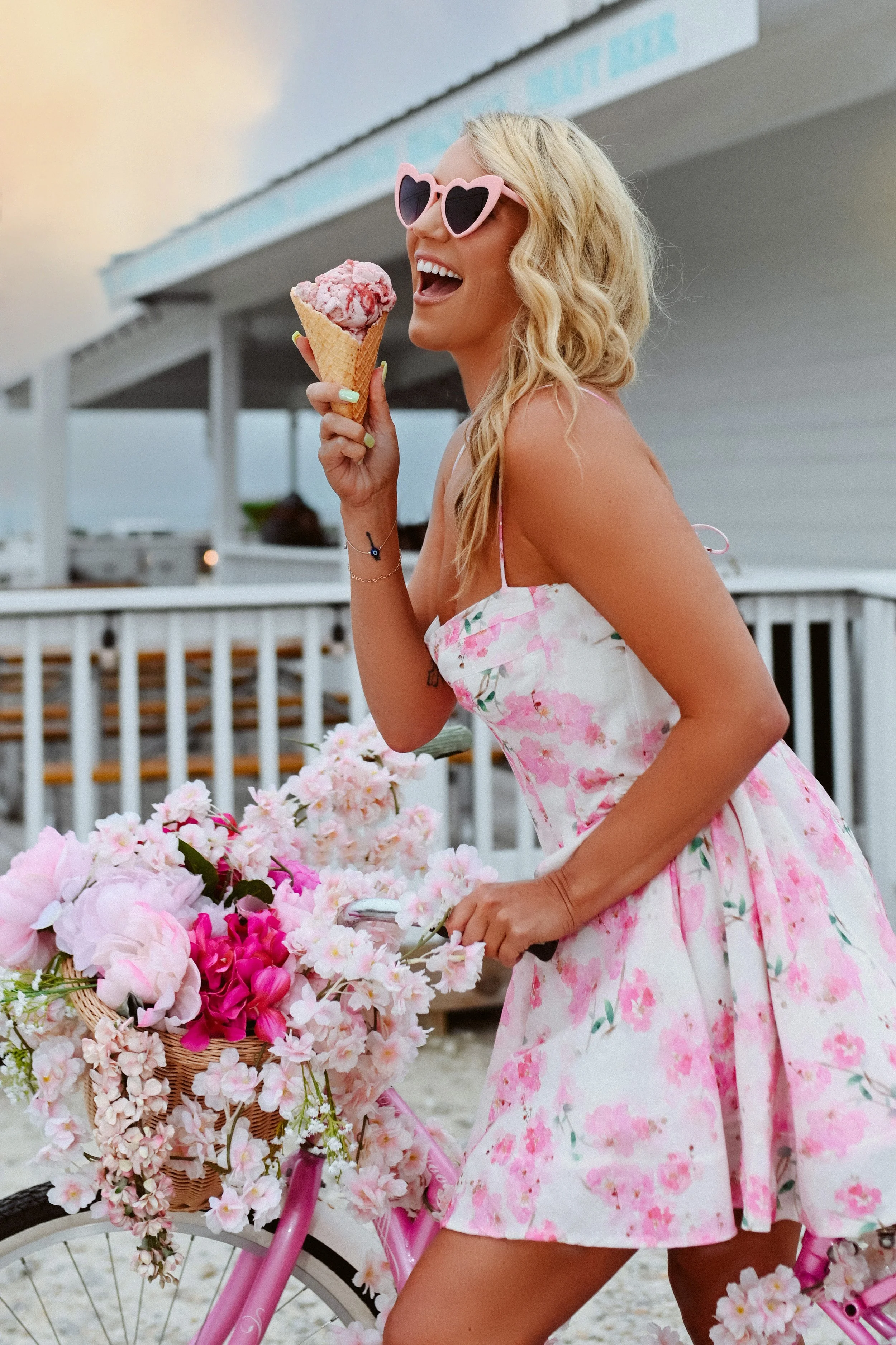 A woman with blonde hair wearing pink heart-shaped sunglasses and a pink floral dress, smiling and holding an ice cream cone, standing next to a pink bicycle decorated with pink flowers.