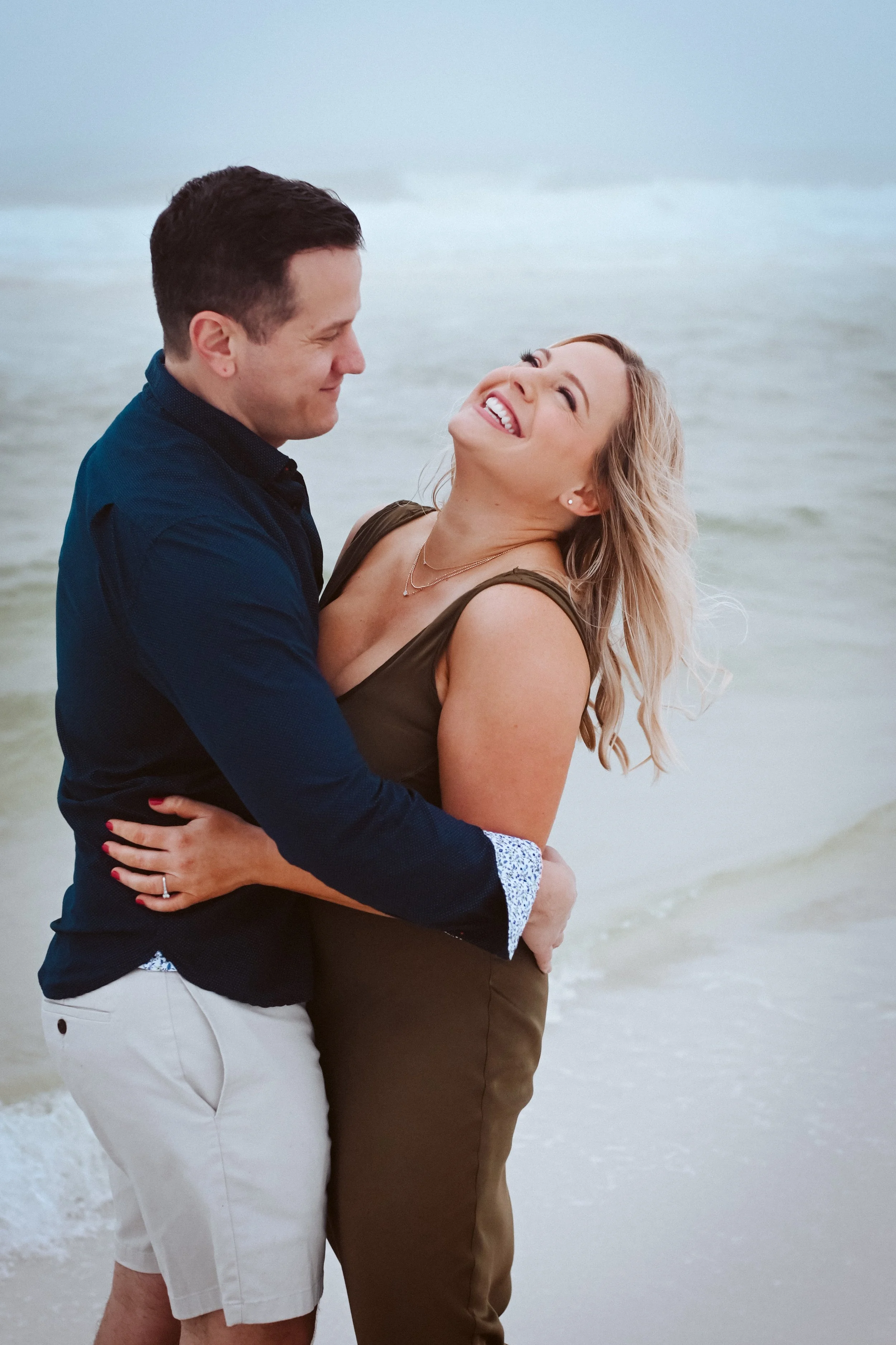 A couple embracing and smiling at each other on a beach with the ocean in the background.