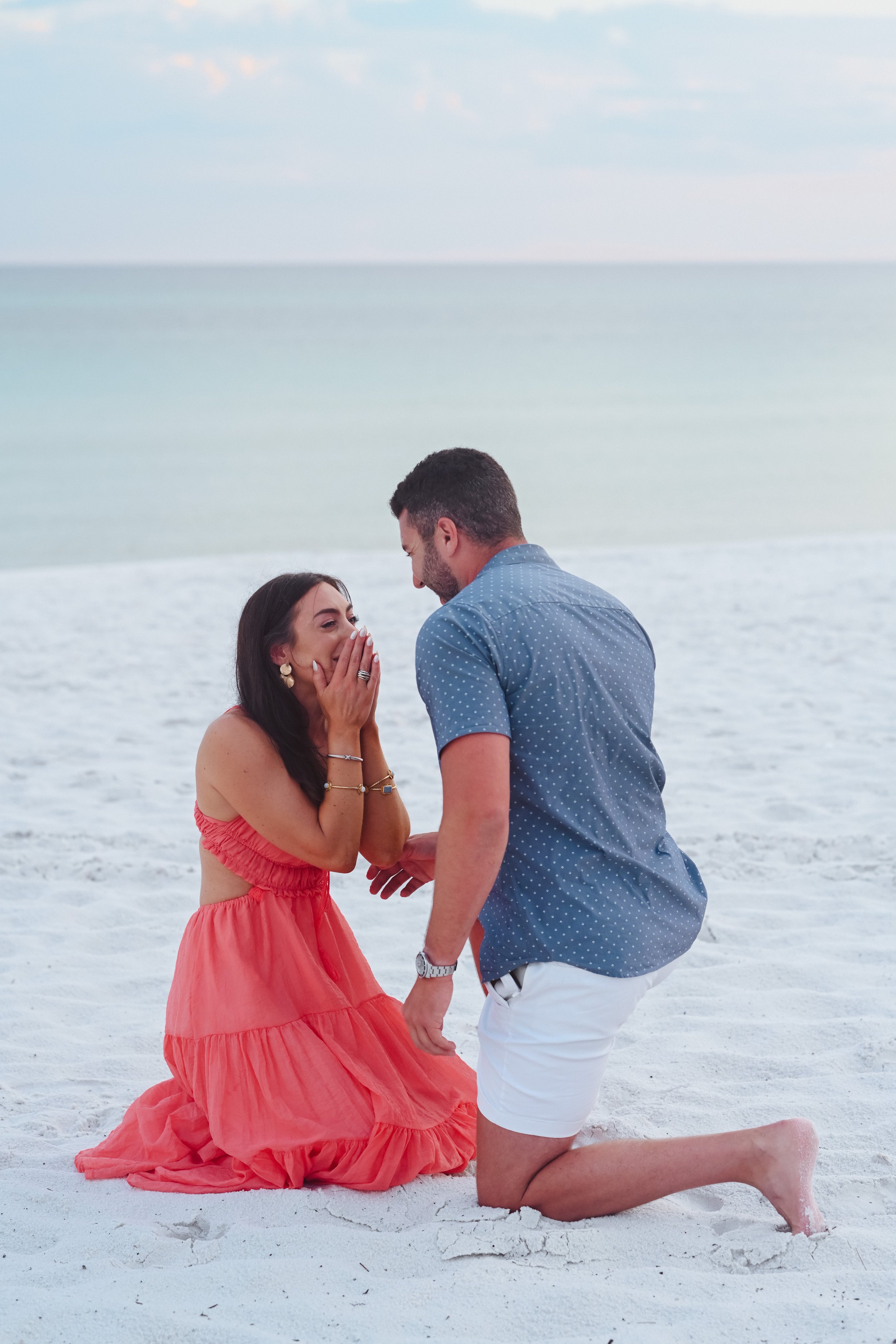 A man on one knee proposing to a woman on a beach, with the woman expressing surprise and joy.