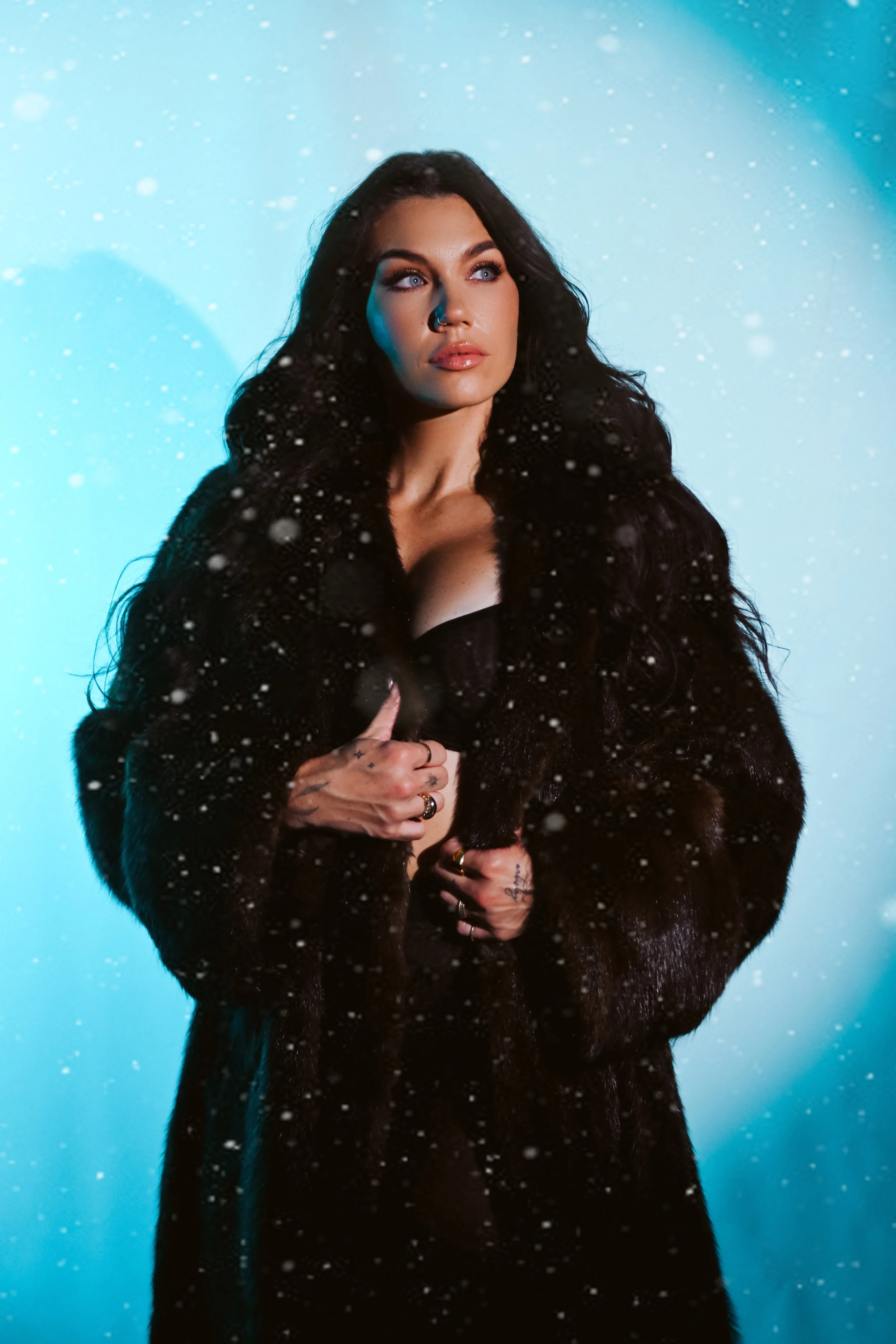 A woman with long dark hair wearing a black fur coat poses in front of a blue background with snow falling around her.