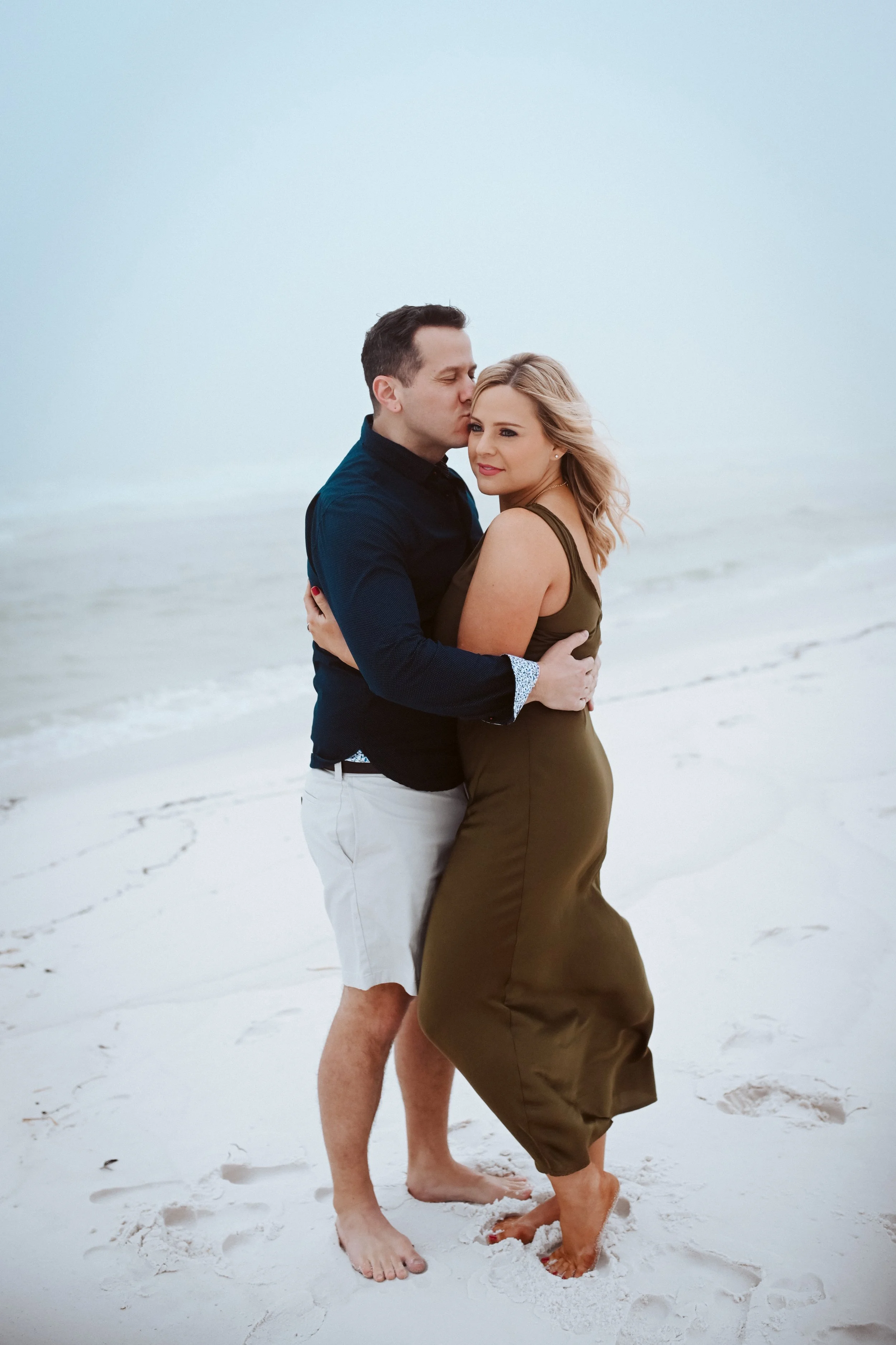A couple embracing on the beach with ocean in the background, the man kissing the woman's temple, both barefoot.