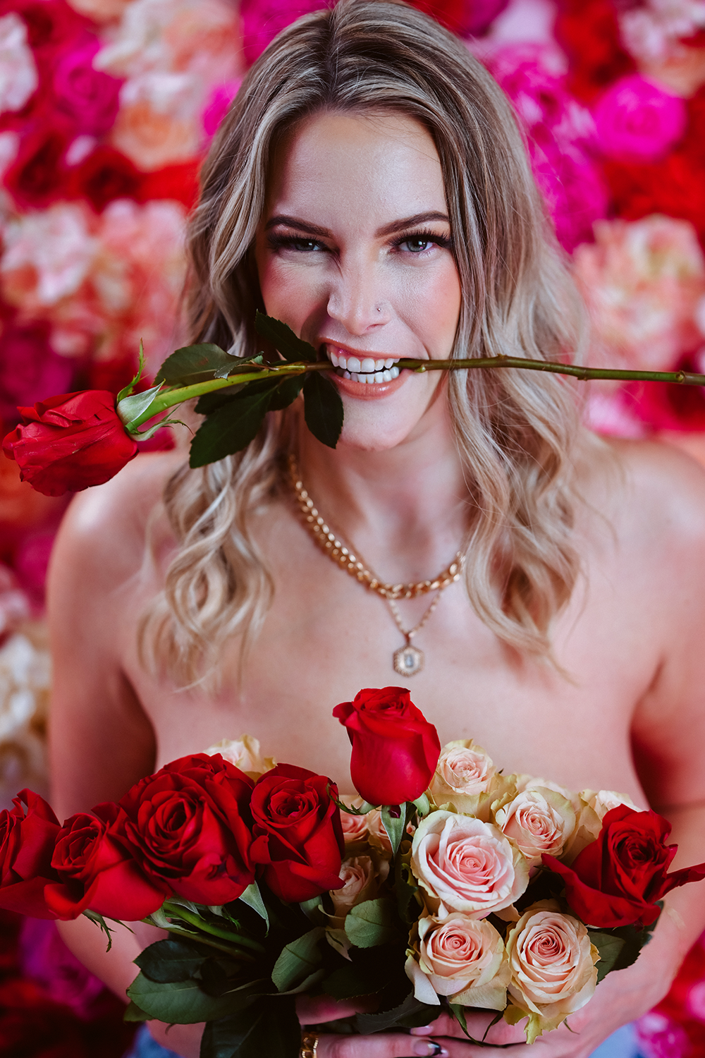 A woman with wavy blonde hair holding a bouquet of red and light pink roses, with a single rose in her mouth, against a background of pink and red flowers, smiling playfully.