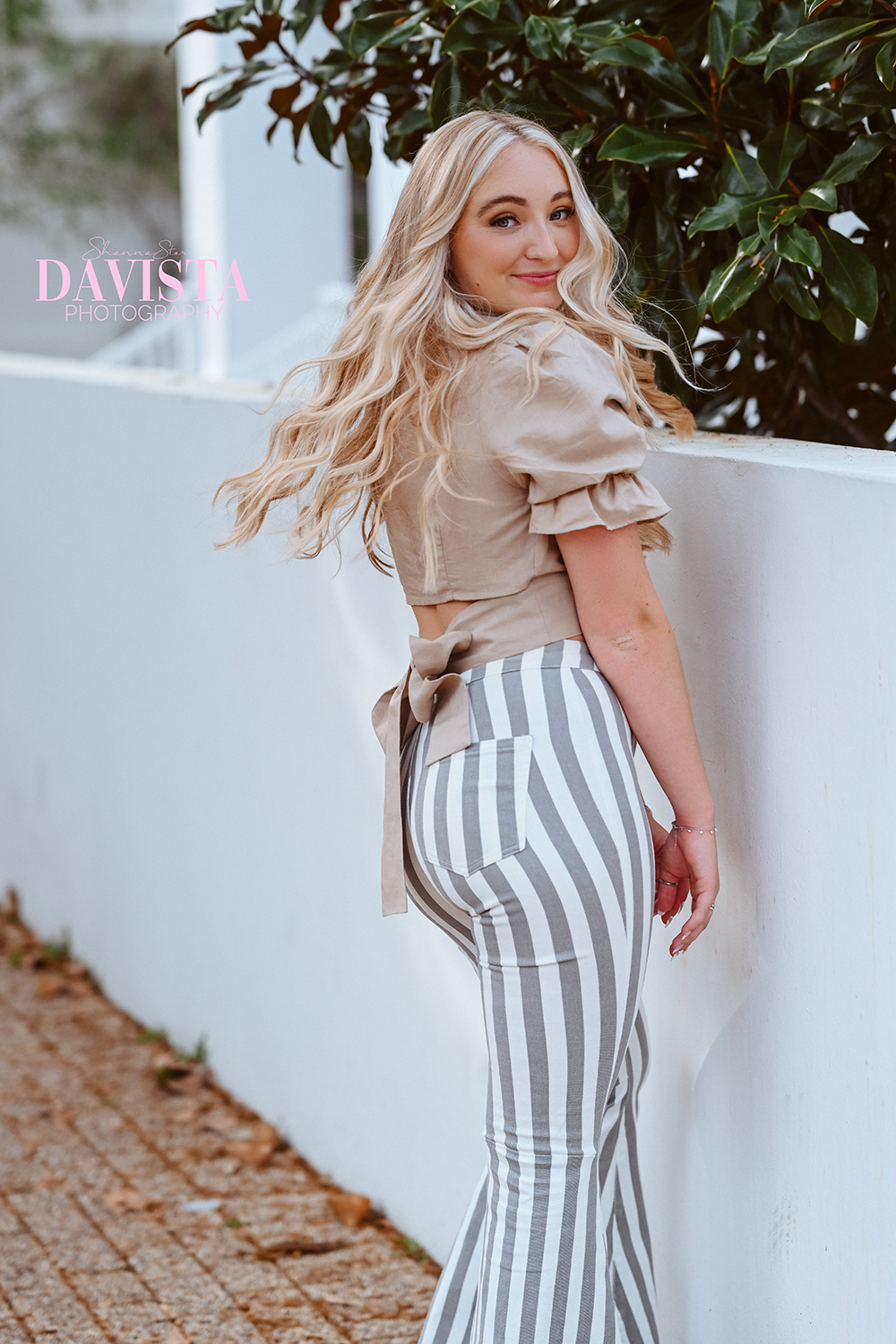 A young woman with long blonde hair and a light makeup look is smiling at the camera while standing outdoors next to a white wall and green bushes. She is wearing a beige puff-sleeve crop top and high-waisted striped pants with vertical gray and whit
