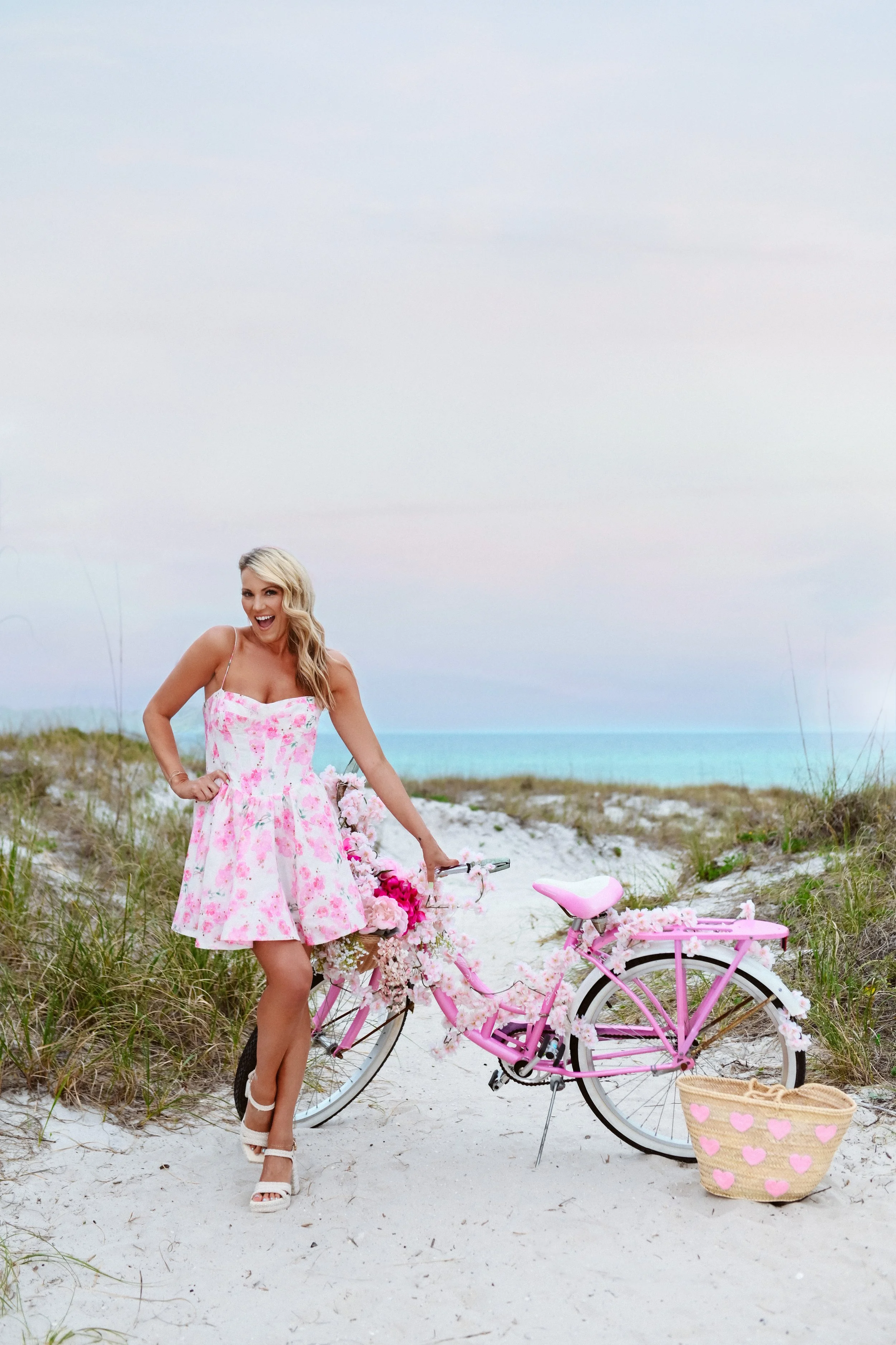A woman in a pink floral dress standing next to a pink bicycle decorated with flowers, on a sandy beach with grass and the ocean in the background.