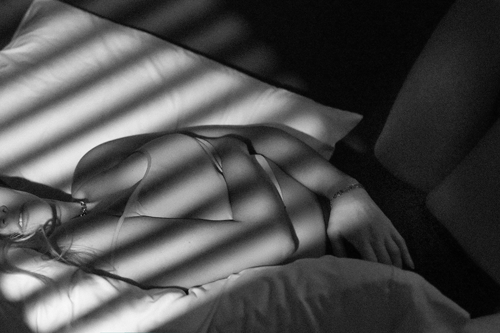 A woman lying on a bed, partially covered by shadows cast by window blinds, wearing lingerie and jewelry.