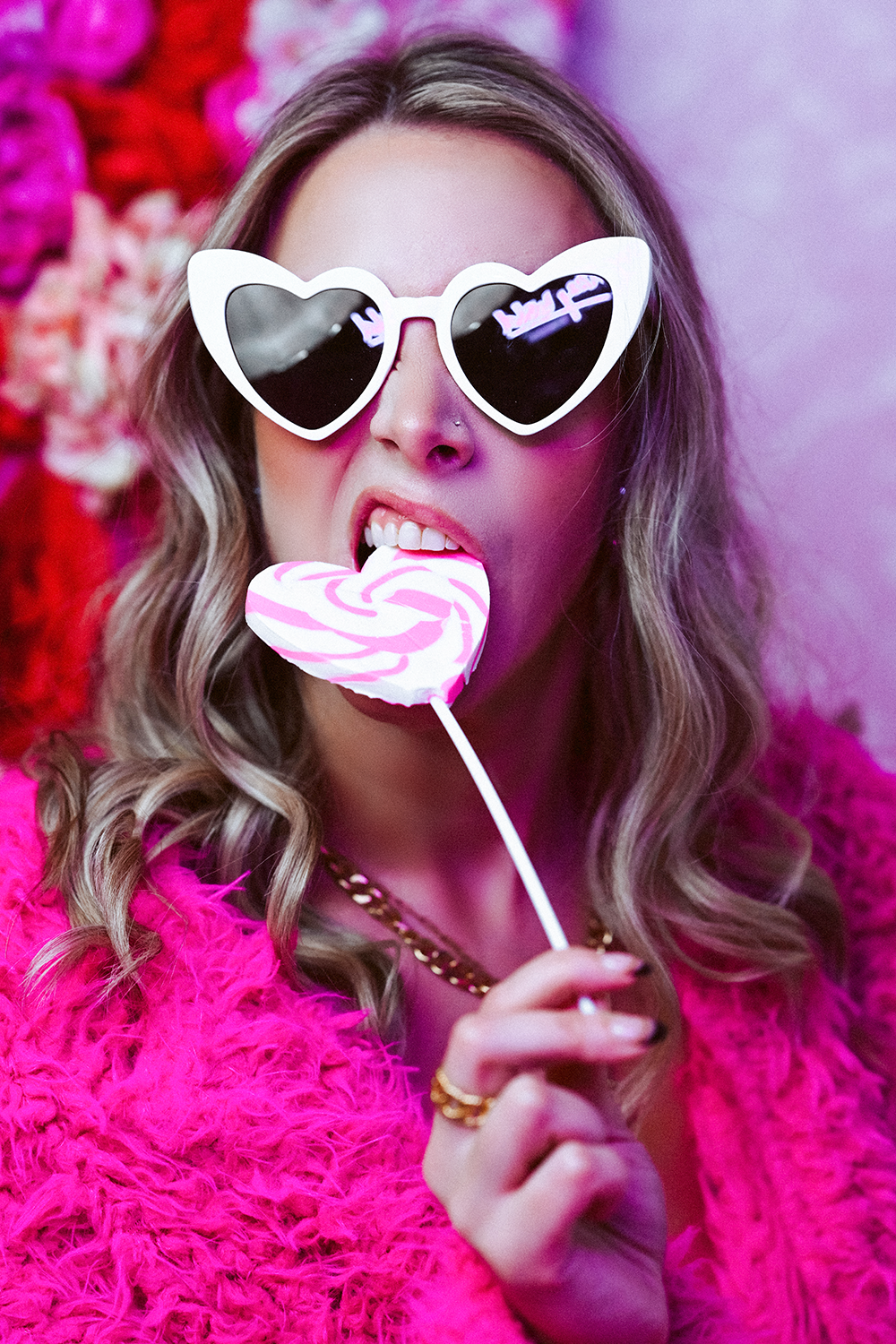 A woman with wavy hair, wearing heart-shaped sunglasses and a pink fuzzy coat, is licking a heart-shaped pink and white lollipop.