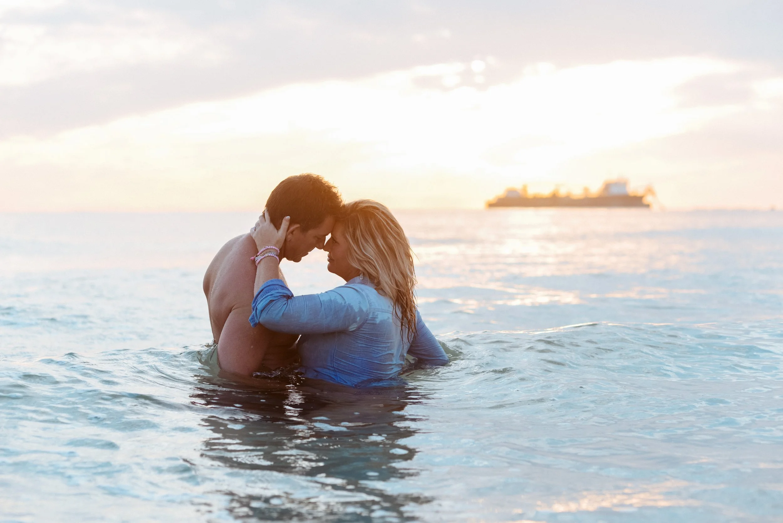 A couple in the ocean embraces at sunset, their foreheads touching, with a distant cargo ship on the horizon.