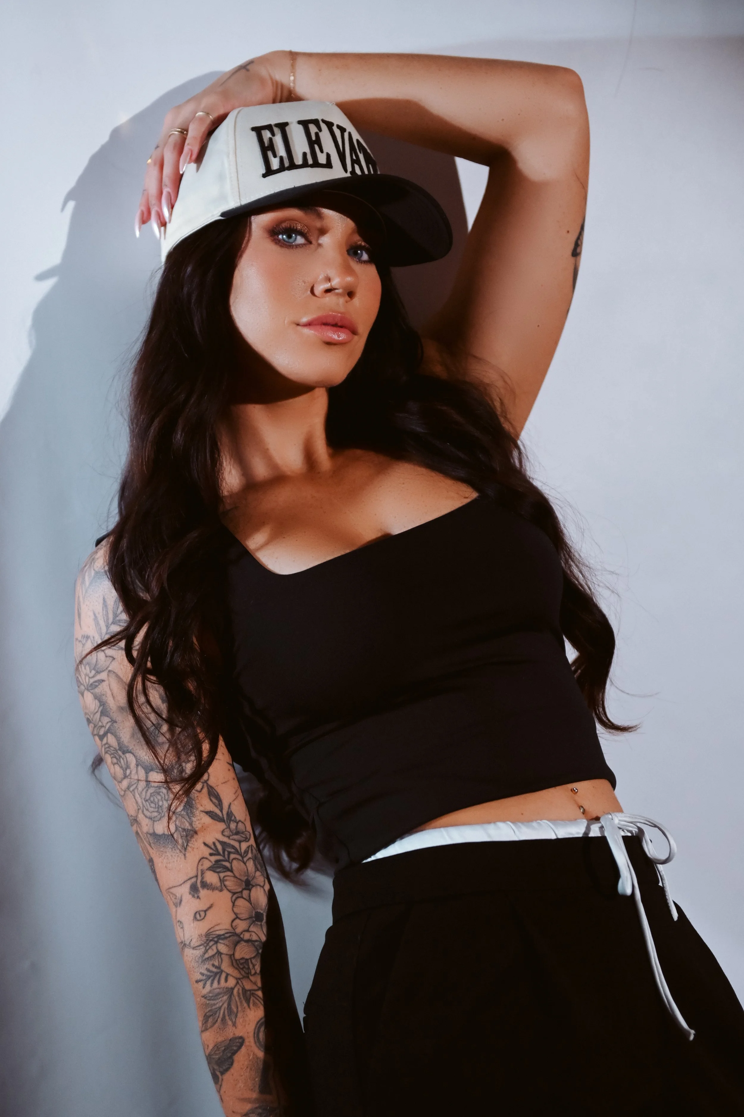 A woman with long dark hair, wearing a black tank top, black pants with a white drawstring, a white and black cap that says 'ELEVAGE,' and tattoos on her left arm, posing against a plain wall.