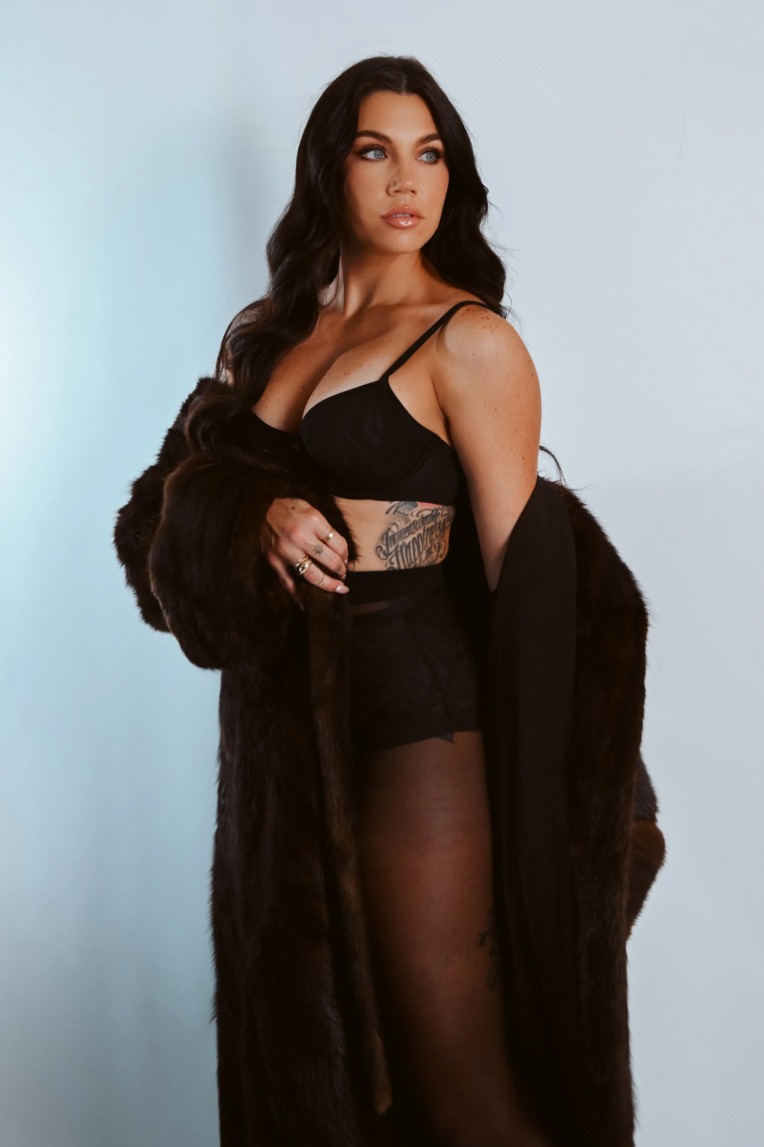 A woman with long dark hair posing, wearing a black bra, black lace shorts, sheer tights, and a dark fur coat draped around her shoulders, with tattoos visible on her stomach and thigh.