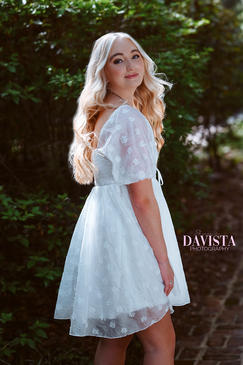 A young woman in a white, embroidered, short-sleeved dress, standing outdoors with green foliage in the background, looking at the camera with a slight smile.