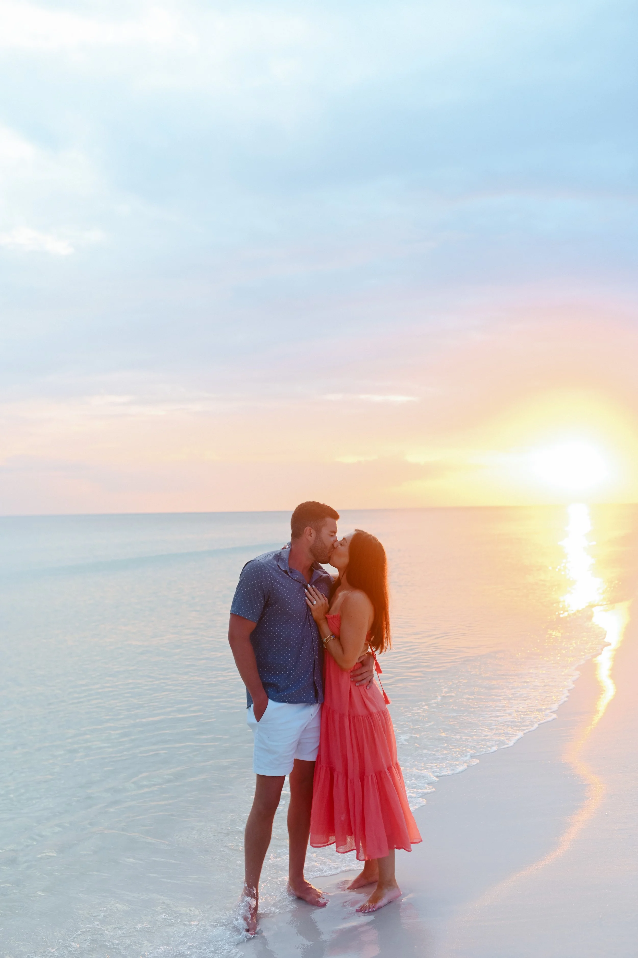 A young couple sharing a kiss on the beach at sunset, with the ocean and colorful sky in the background.