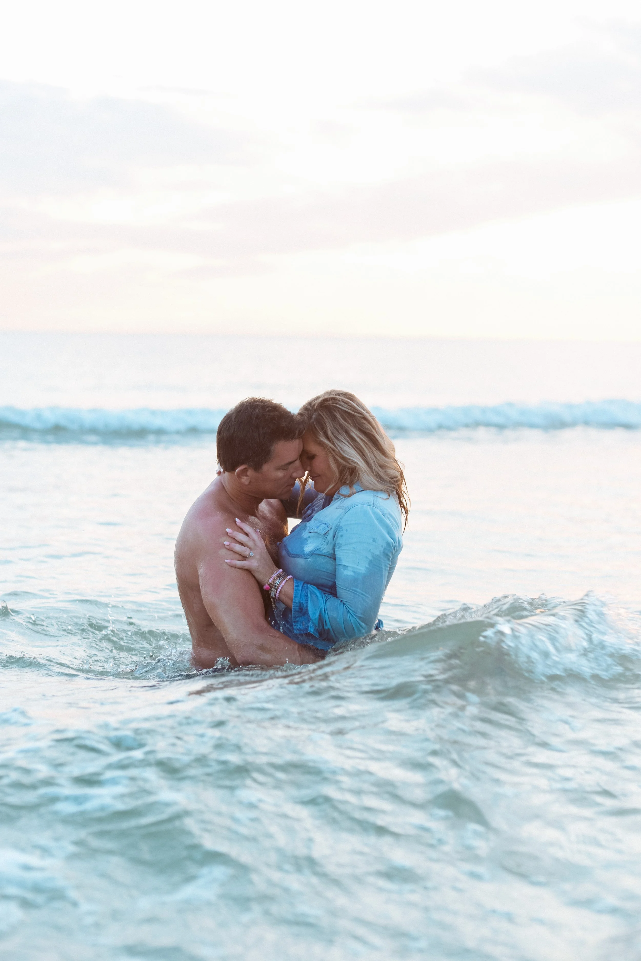 A couple embracing in the ocean during sunset, with waves around them and a cloudy sky above.