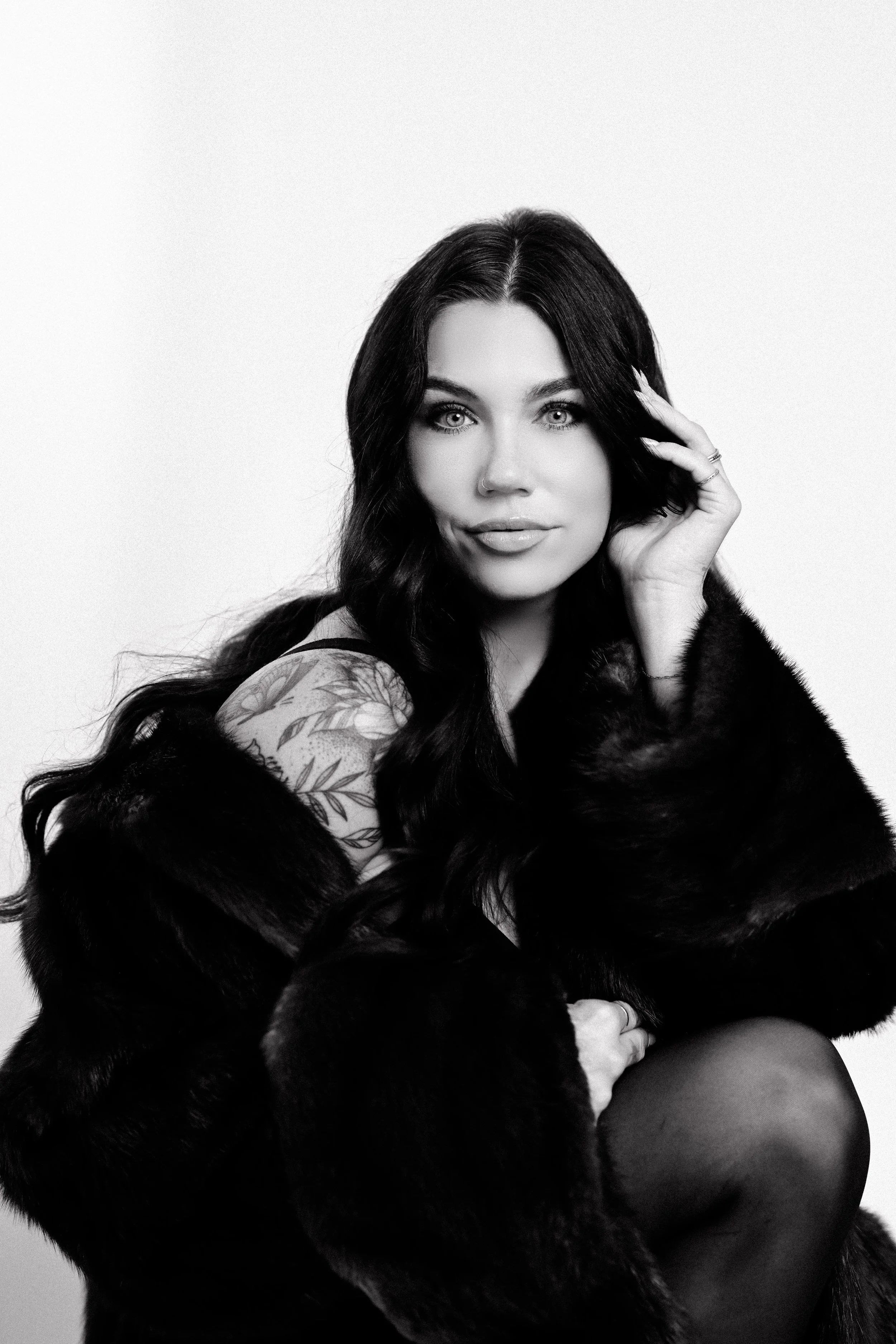 Black and white portrait of a woman with long dark hair, wearing a fur coat and a tattoo on her shoulder, looking at the camera.