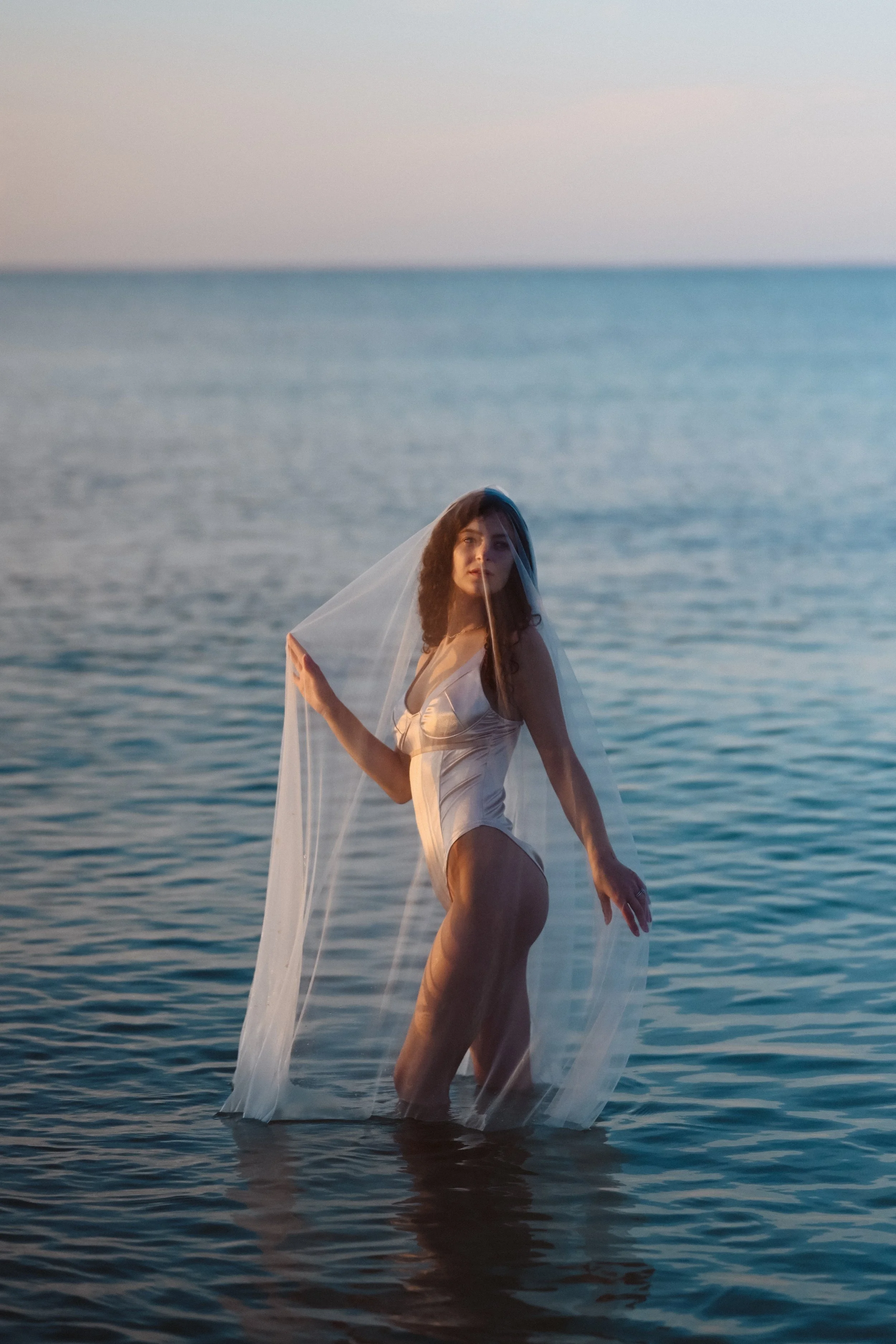 A woman in a wedding dress stands waist-deep in water at sunset or sunrise, holding a veil.