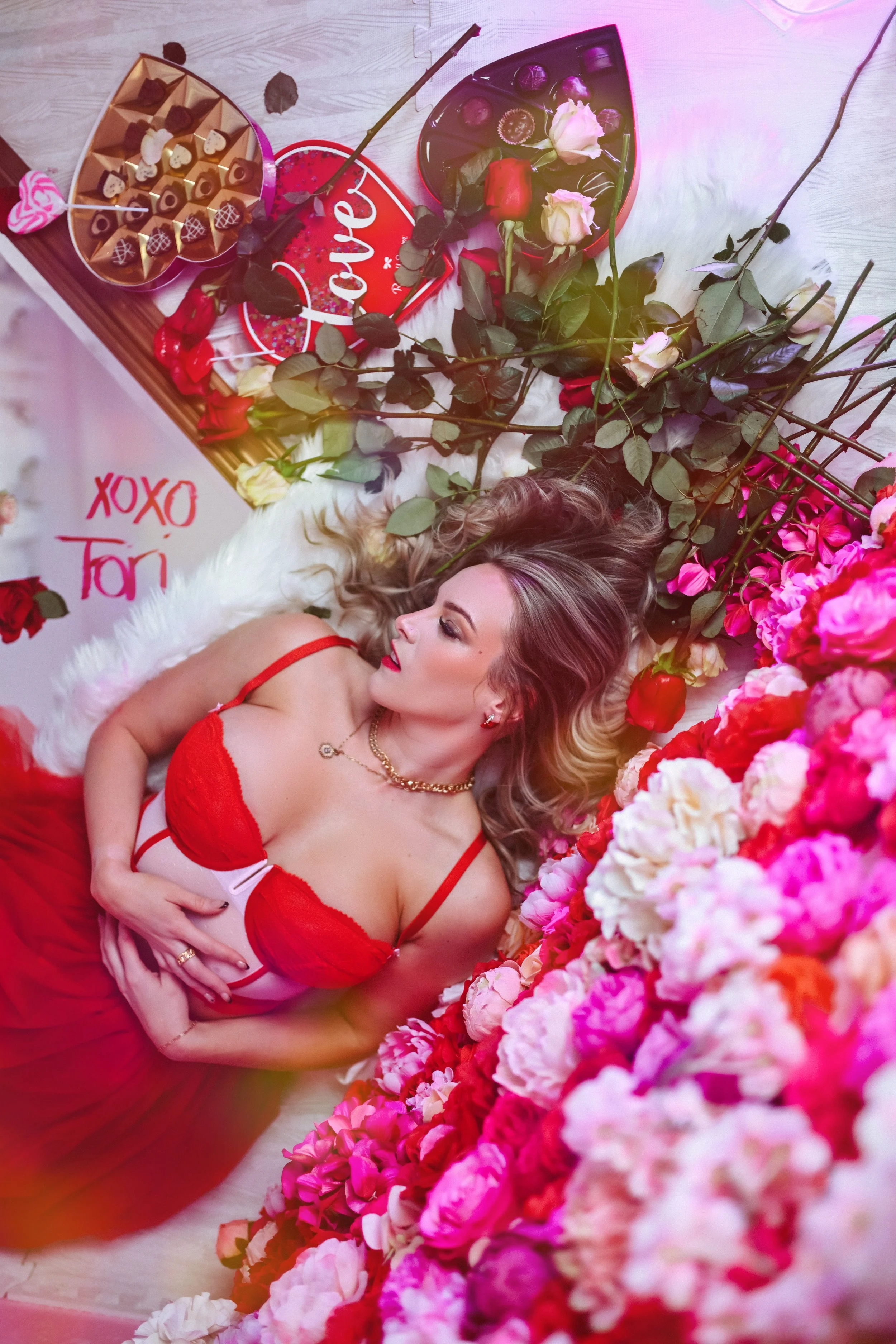 A woman in red lingerie lying on a bed of pink and white flowers, with roses and chocolates surrounding her. There is a sign that says 'XOXO Tori' and a tray with more chocolates and roses, as well as a heart-shaped pillow with the word 'Love' writte