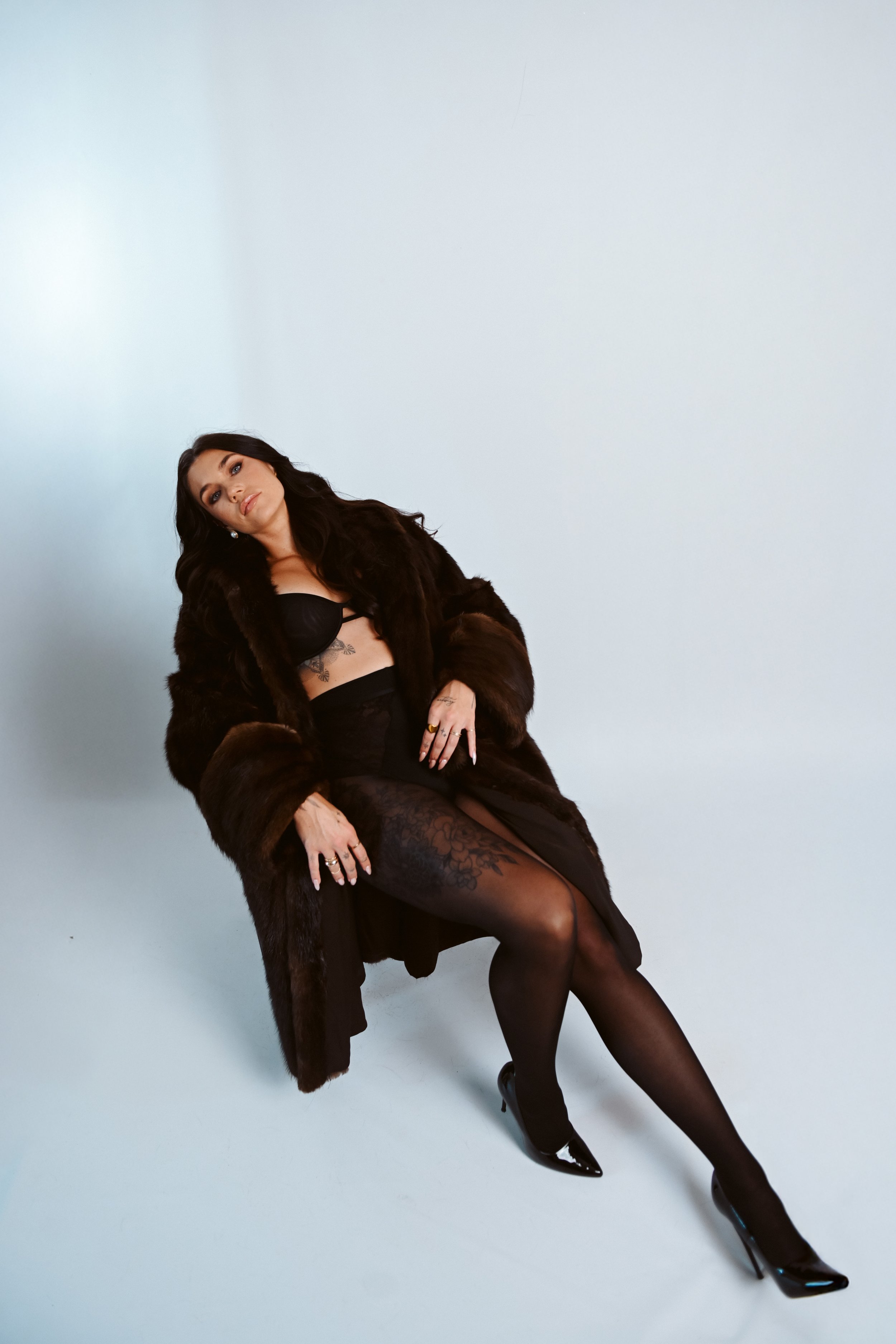 A woman with dark hair wearing a black dress, black stockings, high heels, and a dark fur coat, sitting against a plain light background.