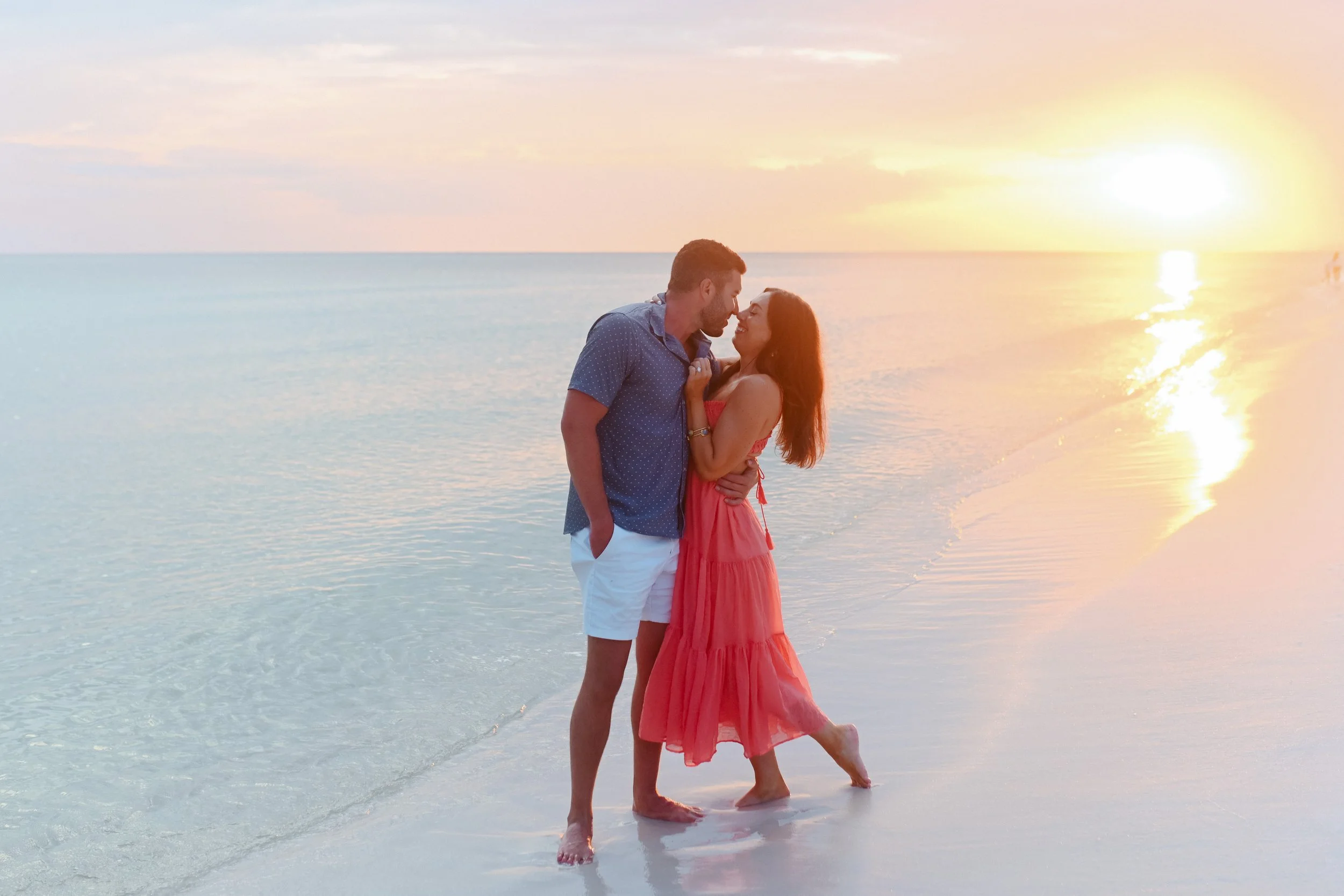 A couple is standing close together on a beach during sunset, with the man in a blue shirt and white shorts and the woman in a coral dress. They are looking at each other lovingly, with the ocean and the setting sun in the background.