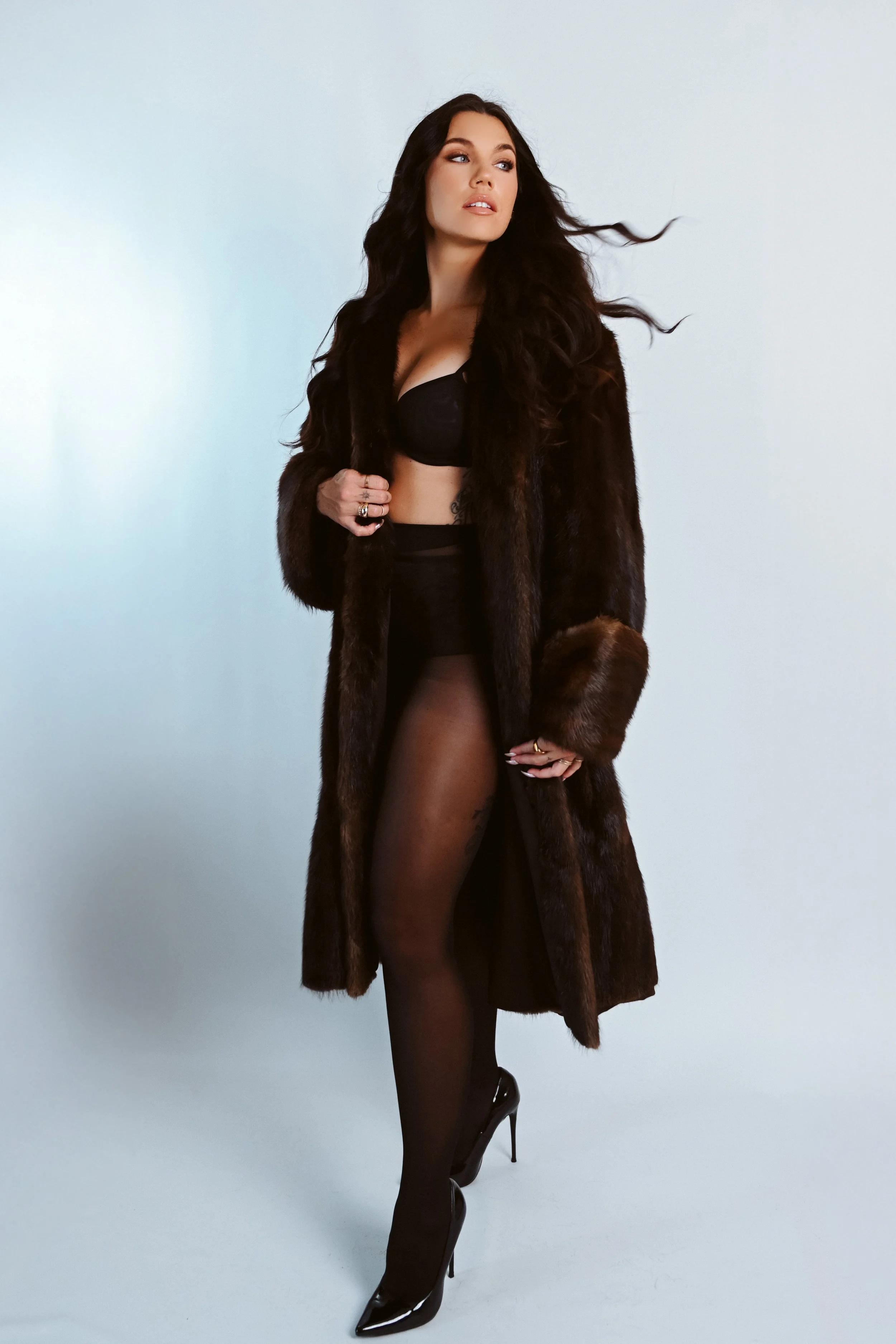 A woman with long dark hair posing in a studio, wearing a long fur coat, black lingerie, sheer black stockings, and shiny black high heels.