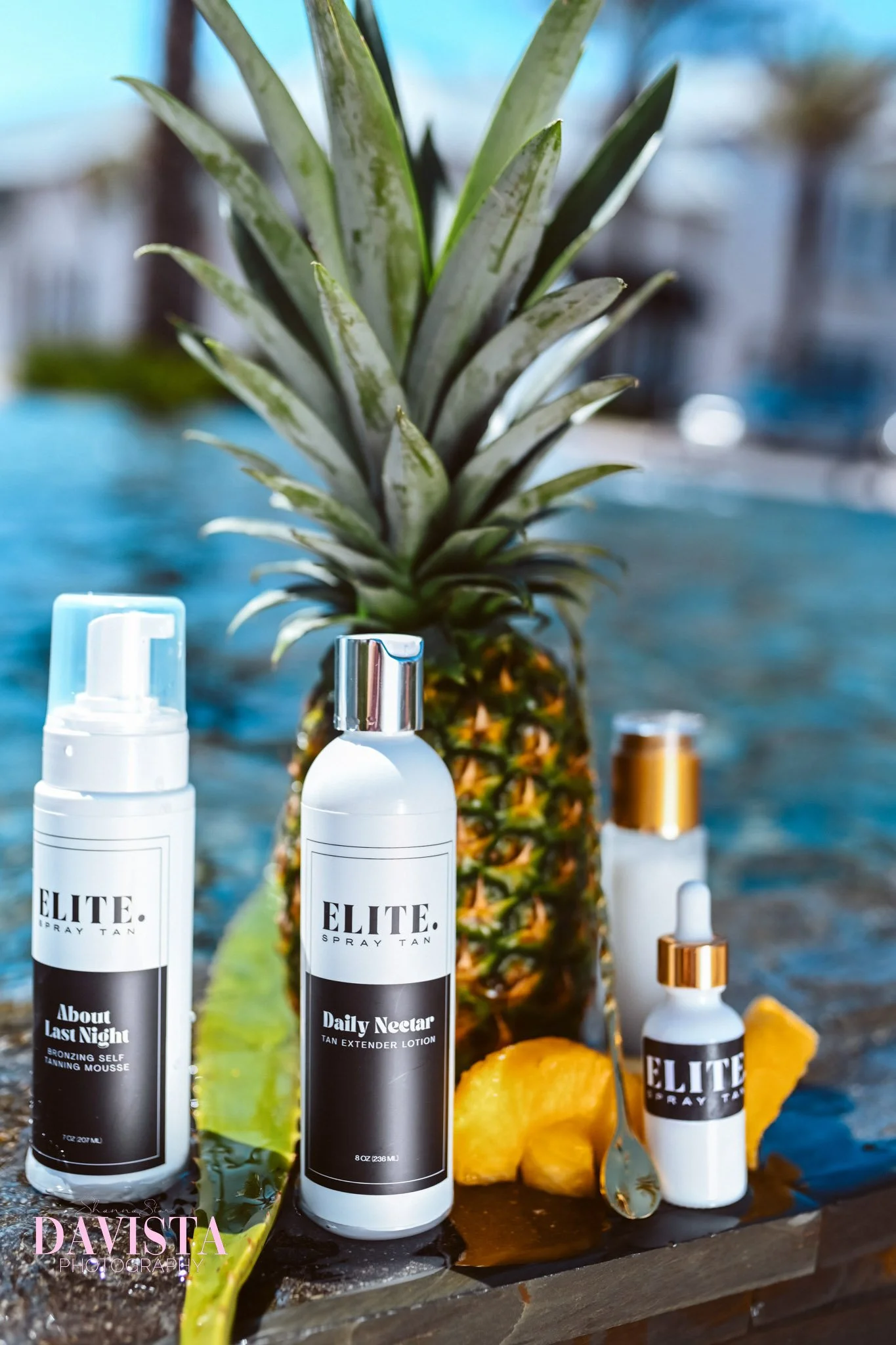 A pineapple surrounded by skincare products including spray tan bottles, a dropper bottle, and lotion, with pineapple slices and green leaves nearby, set outdoors on a wooden surface.