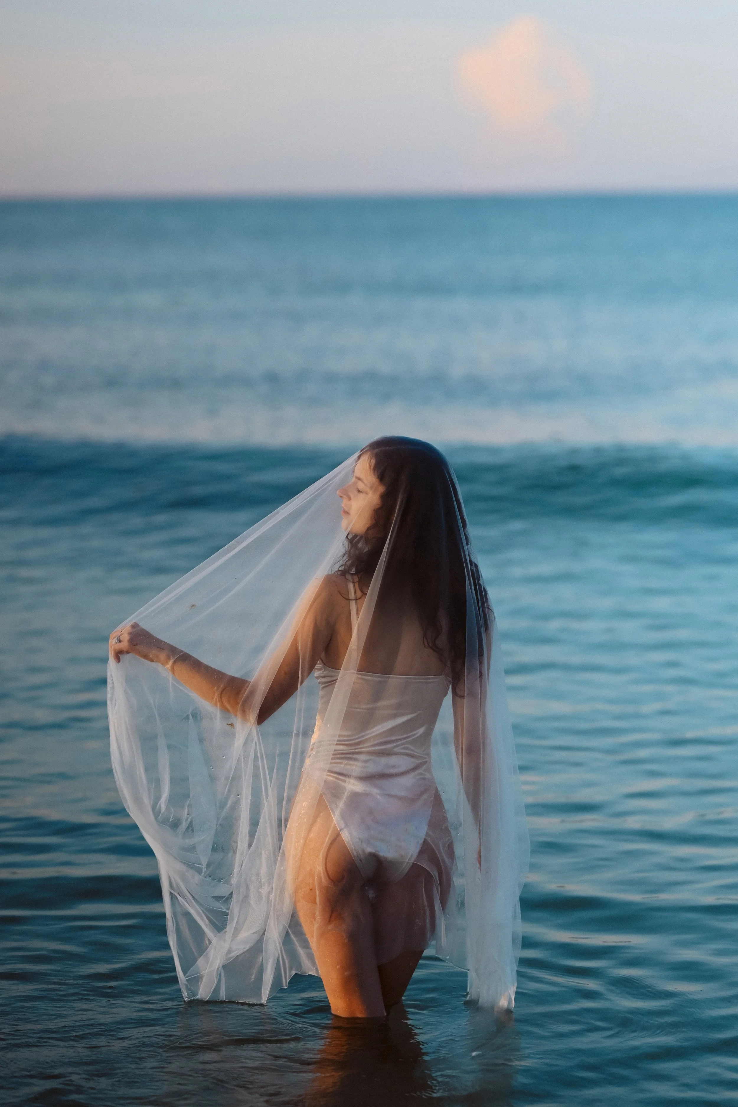 A woman stands in the ocean with water up to her thighs, holding a sheer veil over her head and shoulders, facing sideways with closed eyes, during sunset or sunrise.