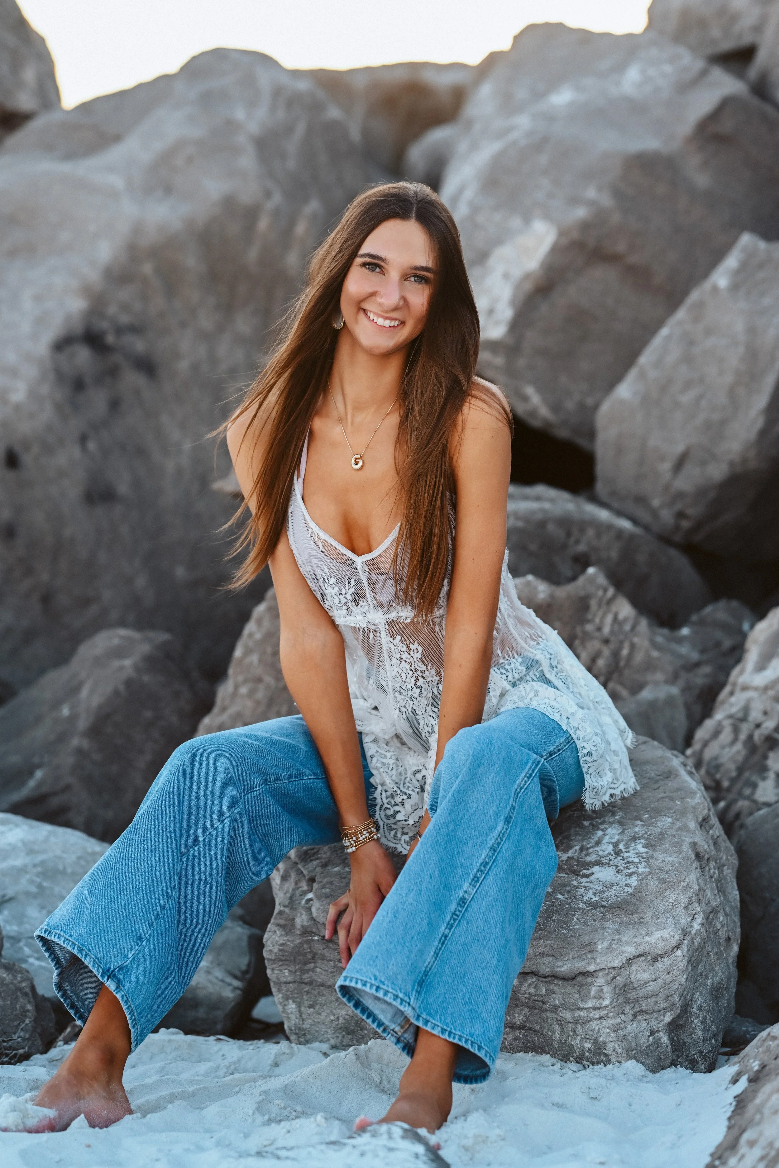 A young woman sitting on a large rock among other rocks at the beach, smiling at the camera, wearing a white lace dress and blue jeans.