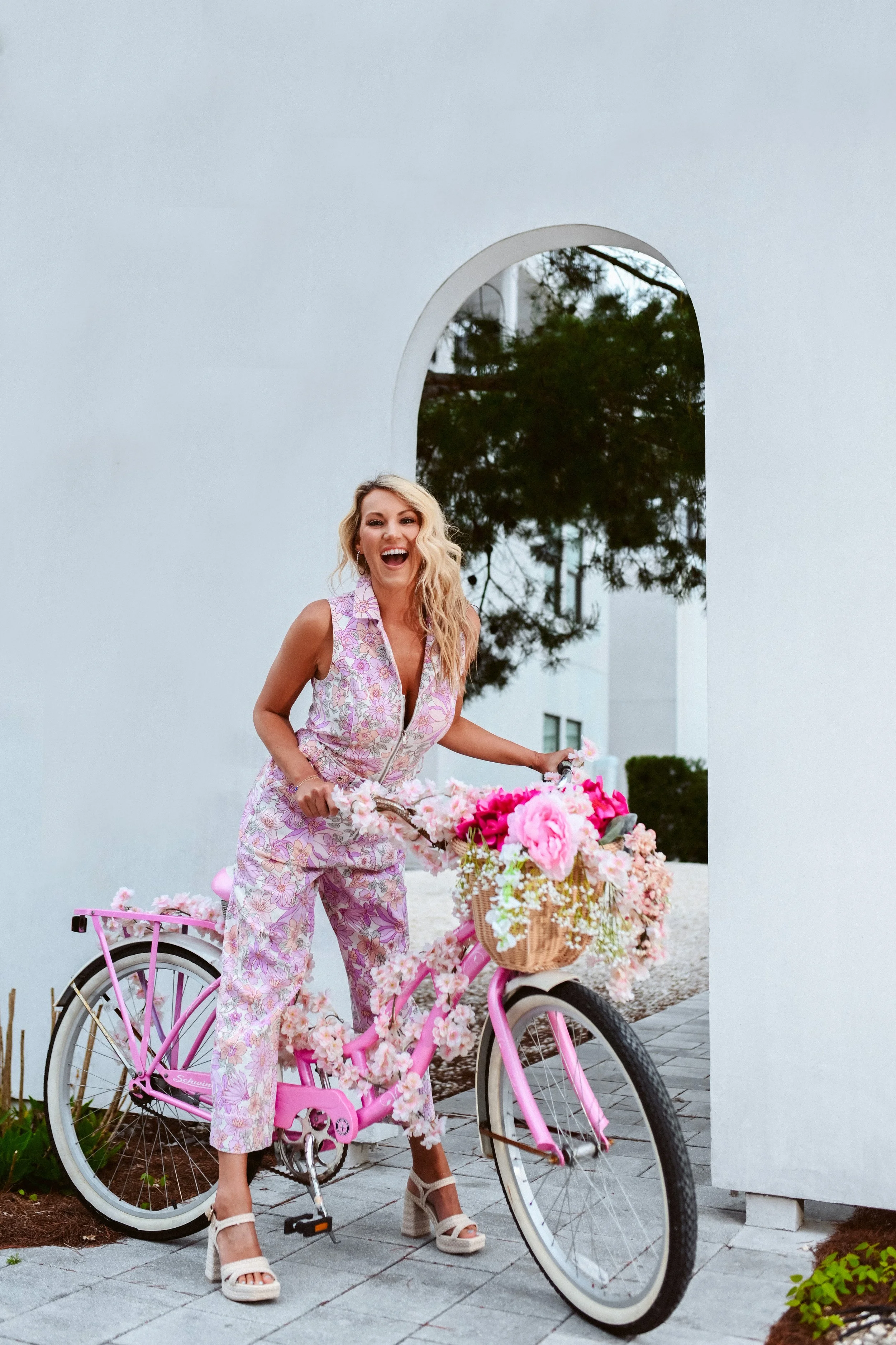 A woman with blonde hair wearing a sleeveless pink floral jumpsuit and white high heels, standing next to a pink bicycle decorated with pink and white flowers. She is laughing happily outdoors in front of a modern white wall with an archway, with gre