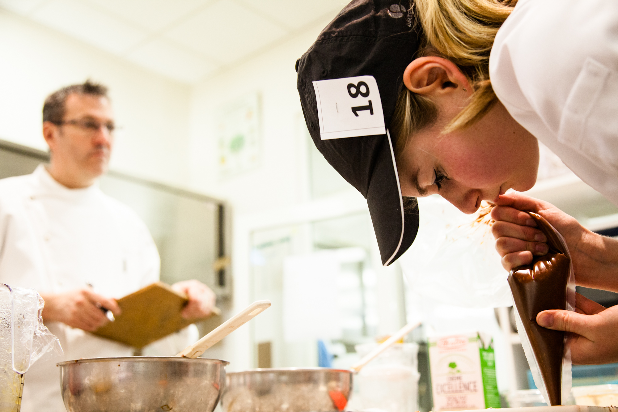  Une candidate poche un entremet pendant qu'un des membres du jury de travail regarde un autre candidat travailler. 