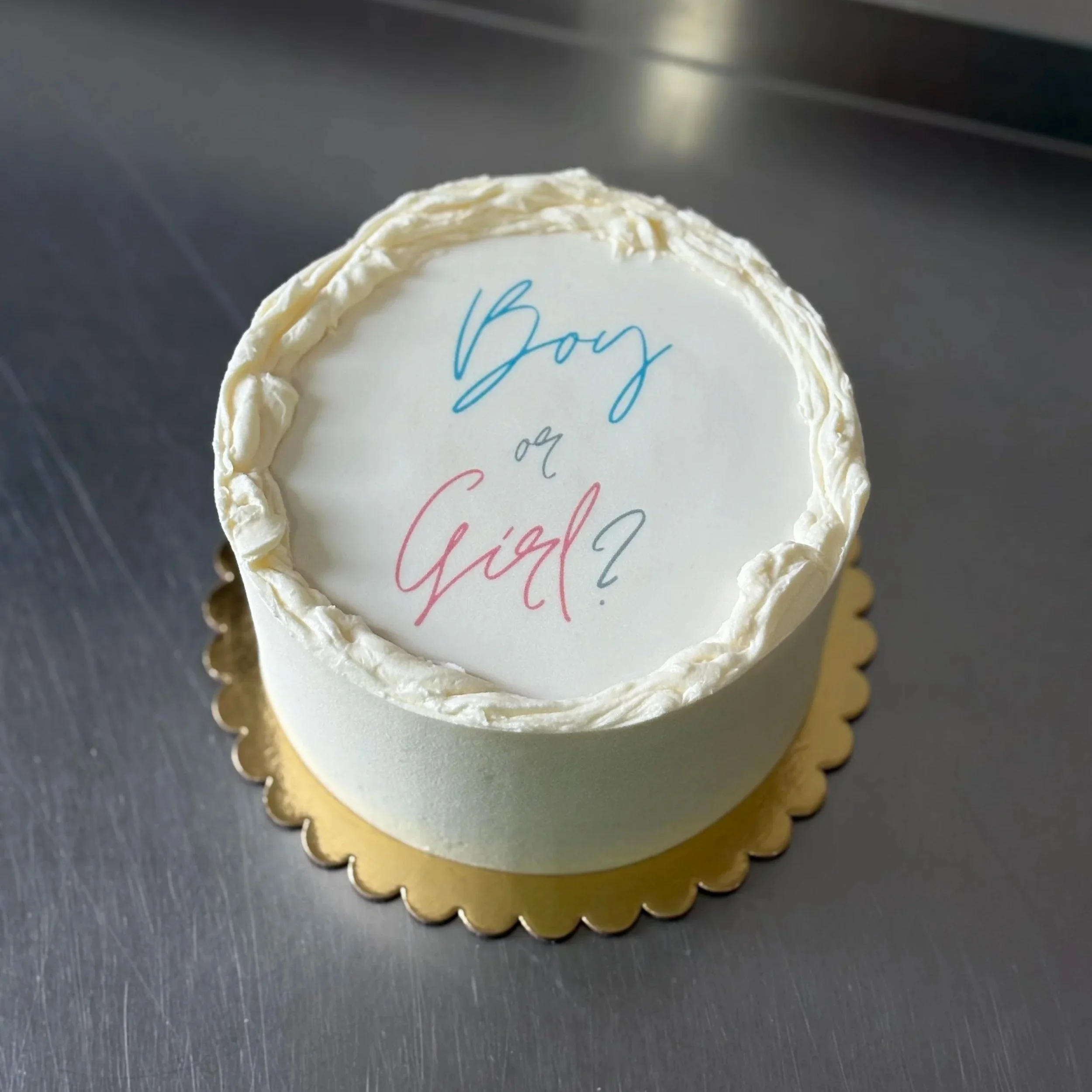 A picture of a vanilla white cake with a rustic edge and boy (in blue) or (in grey) girl (in pink)edible image on top with a grey question mark.