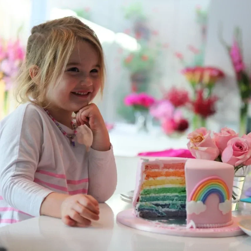A picture of a little girl sitting next to her rainbow layered cake with pink fondant icing and an icing rainbow on the front. A slice is cut out so you can see the rainbow layers.