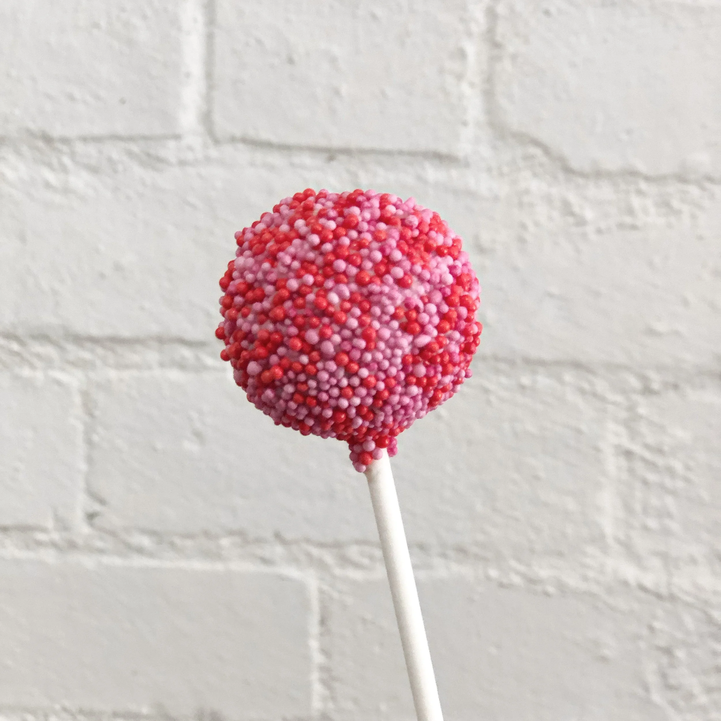 A picture of a cake pop covered in pink and red 100s and 1000s.