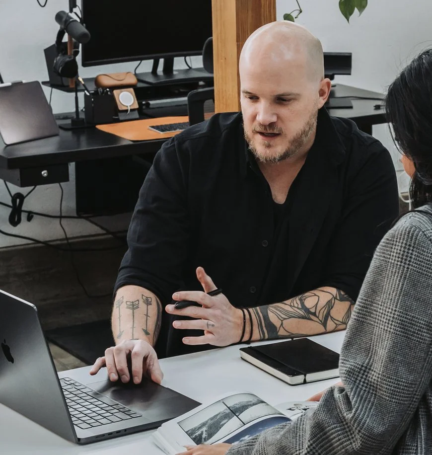 Two people having a discussion at a desk in an office, with a laptop, a notebook, and a magazine. A computer monitor and headphones are visible in the background.