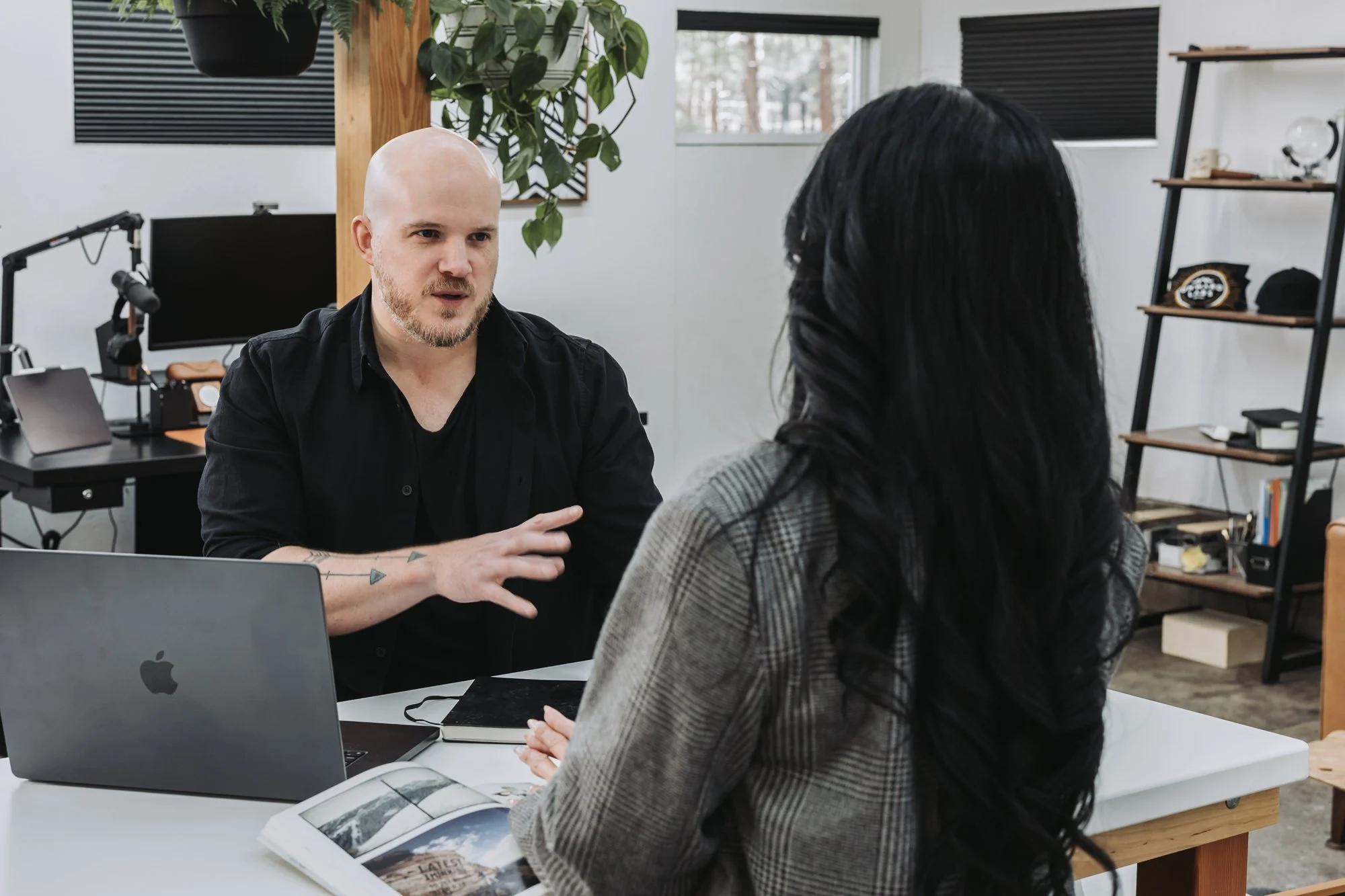 Ryan is talking to a woman with long black hair. They are sitting at a white table in an office with computers and books. The man appears to be explaining or discussing something.